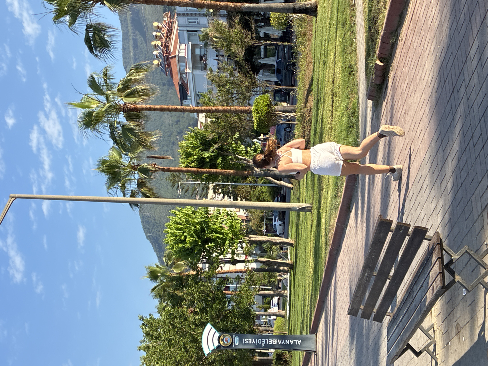 In Alanya, Turkey, a young woman with brown hair, wearing a white sports bra and white shorts, is captured mid-stride as she runs away from the camera on a paved walkway. Her movement is dynamic, with her left leg extended backward and her right leg bent as she propels herself forward. She is wearing white athletic shoes with dark socks. To the left of the walkway, a dark wooden bench with metal supports is positioned on a patch of grass. A tall, light-colored pole with a wifi symbol and text reading "ALANYA BELEDIYESI" stands prominently near the bench. The background reveals a lush green park area with various trees, including several tall palm trees. Beyond the park, a multi-story building with a red-tiled roof and solar water heaters is visible, situated against a backdrop of a lush, green mountain. The sky above is a clear blue with scattered white clouds, and the sun casts distinct shadows on the walkway, indicating it is likely morning or late afternoon. Cars are parked and driving on a road visible behind the park, suggesting a public urban setting.