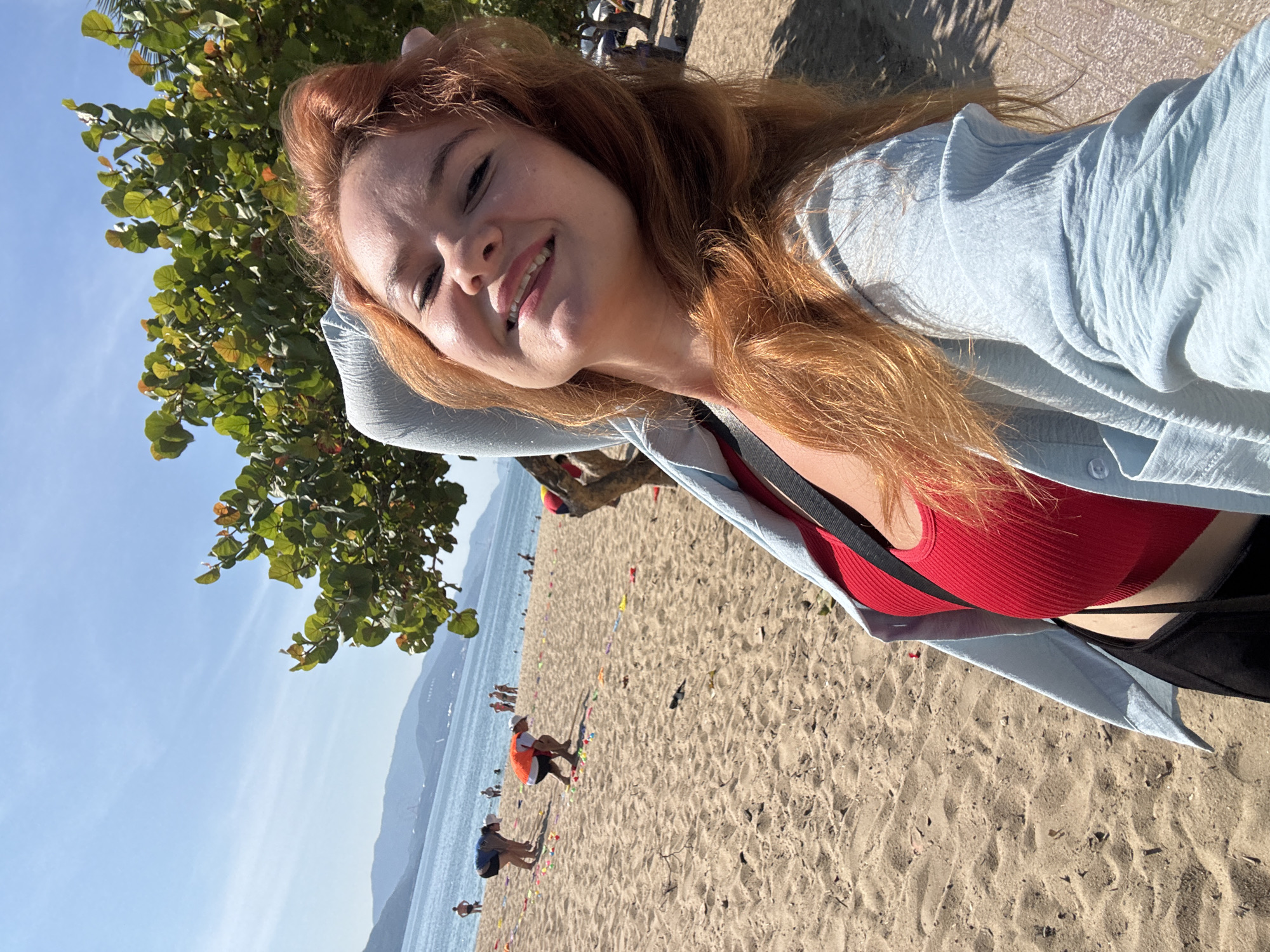 A young Caucasian woman with long, reddish-blonde hair and fair skin takes a selfie on a sandy beach. She is wearing a red, ribbed tank top and a light blue, open button-up shirt. Her right hand is behind her head, and she is smiling at the camera. In the background, the ocean stretches out to the horizon, with a line of mountains visible in the distance. The beach is populated with several people, some of whom are bending down, possibly participating in an activity. A tree with green leaves is to the right of the woman, casting shadows on the ground. The sand appears light brown and is strewn with small debris. The sky is clear and blue. The scene is set in Nha Trang Ward, Vietnam.