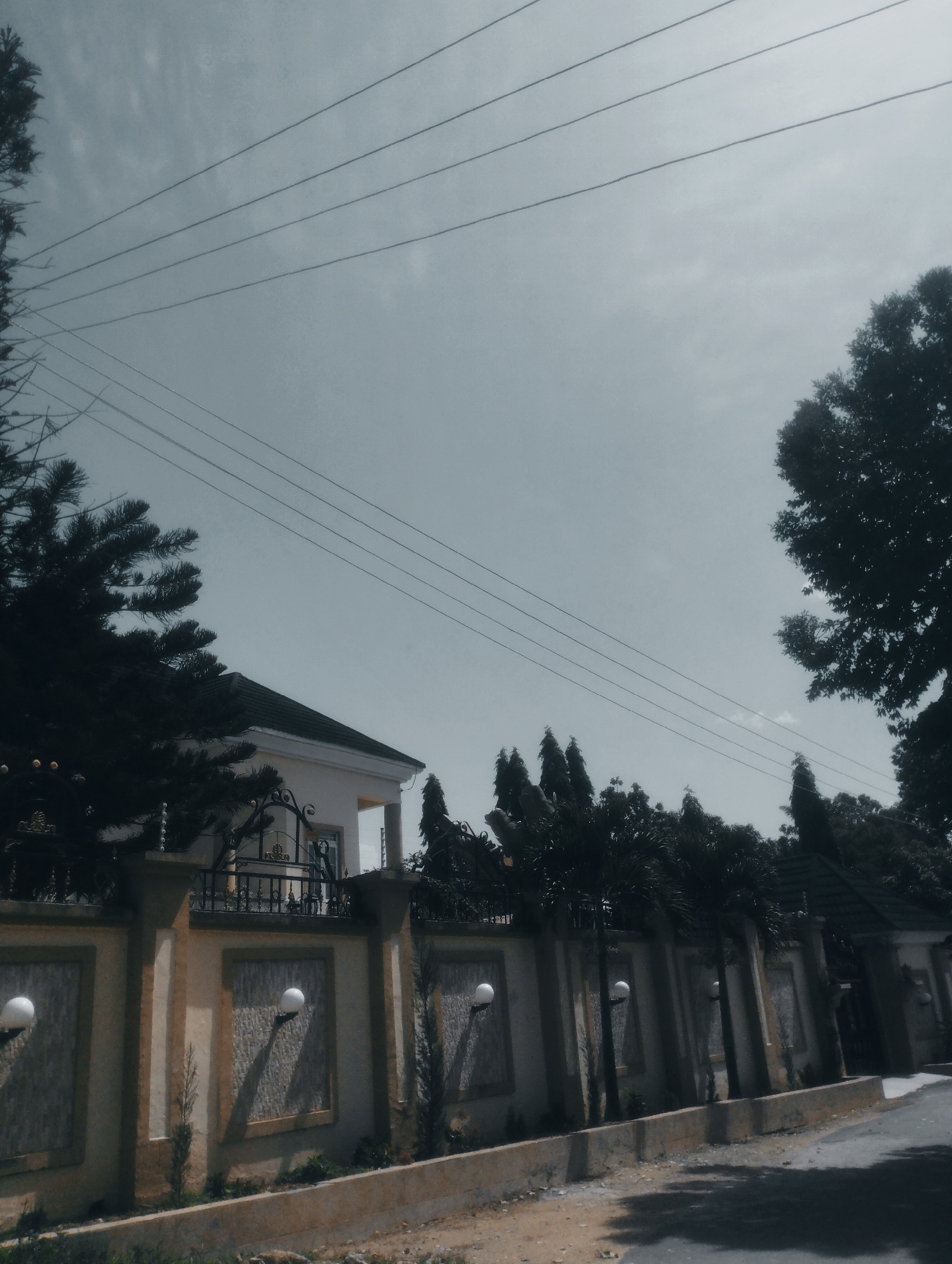 The image captures a view of a residential area in Jos, Nigeria. In the foreground, a concrete curb lines a dusty path adjacent to a paved road. Beyond the path, a decorative wall with textured panels and embedded spherical lights stretches across the scene. The wall is topped with a black wrought-iron fence, with a glimpse of a modern white house with a dark-tiled roof visible behind it. Several tall, slender trees and other foliage are planted along the wall, with larger, mature trees framing the property and the sky. Multiple electrical wires are strung across the pale, overcast sky, adding a common urban element to the landscape. The overall impression is of a well-maintained, suburban residence in Jos.