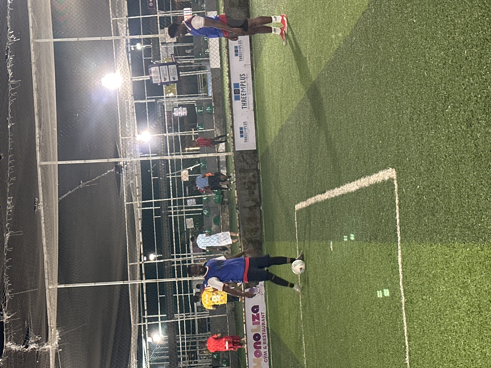 The image shows an outdoor, artificial turf soccer field at night, illuminated by overhead lights. The field has white boundary lines. In the foreground, a Black man wearing a blue vest over a white t-shirt and black shorts with red trim on the sides is playing soccer. He is looking towards the camera and has a soccer ball at his feet. In the midground, several other people are visible on the field, some of whom appear to be playing soccer. To the left, a sign reads "MONOLIZA GYM & RESTAURANT". In the background, there are fences and buildings. The image appears to be taken in Abuja, Nigeria, given the "MONOLIZA GYM & RESTAURANT" sign, which is located in Abuja.