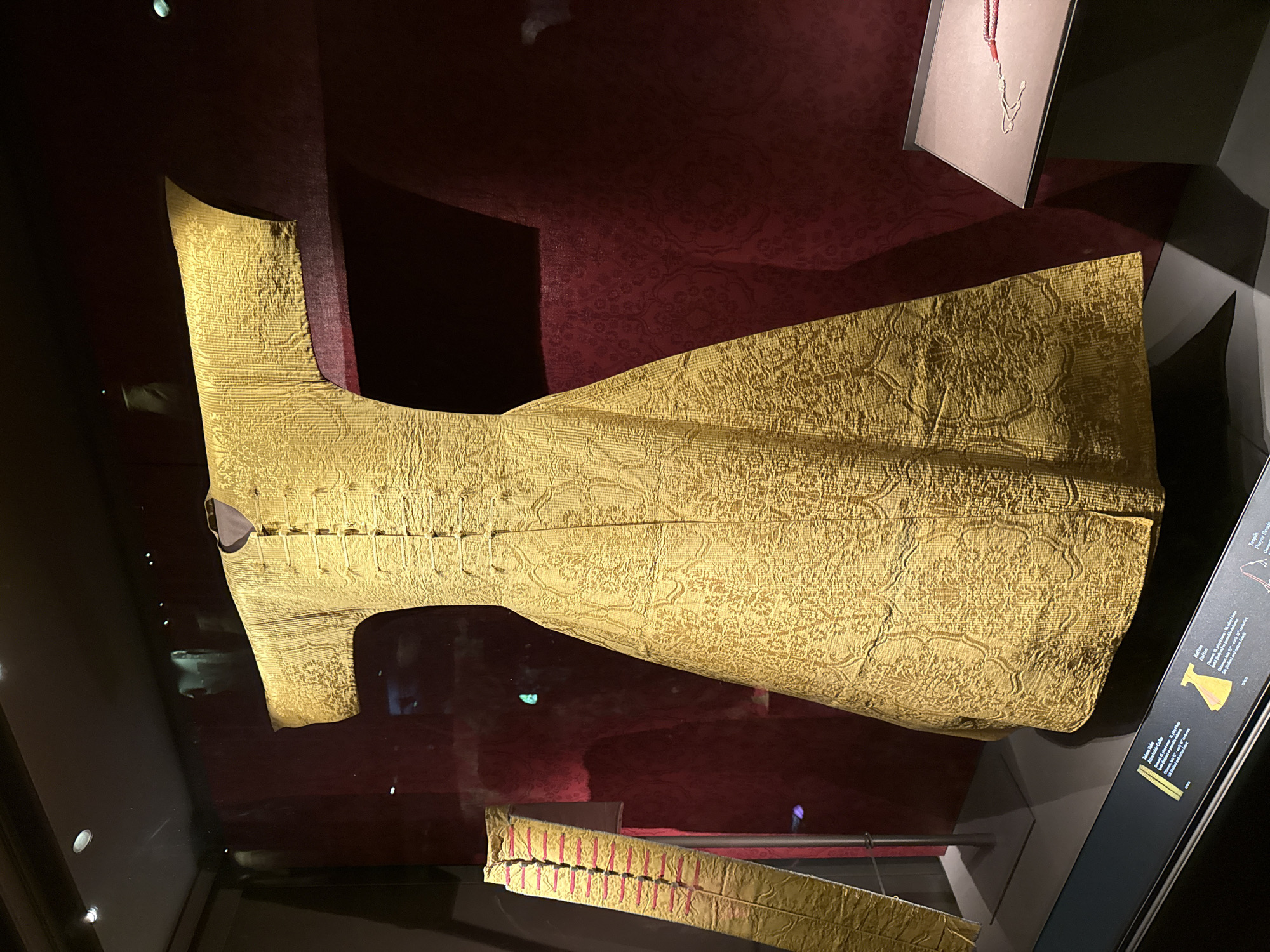 This image shows a display inside a museum, featuring historical garments. The central focus is a long, gold brocade robe with intricate patterns. The robe has wide sleeves and a V-neckline with a row of decorative buttons down the front. To the left of the robe, a portion of a yellow sash with red trim is visible, leaning against a stand. In the background, a dark red fabric with a patterned texture is displayed. To the right of the robe, a small display case contains what appears to be prayer beads with a silver pendant. The museum display is behind glass, and reflections of lights and possibly people are visible on the glass surface. The overall impression is a museum exhibition of historical textiles. The context of Istanbul, Turkey is not explicitly visible in the image, but the presence of such historical garments suggests a location like the Topkapi Palace Museum or another major museum in Istanbul.