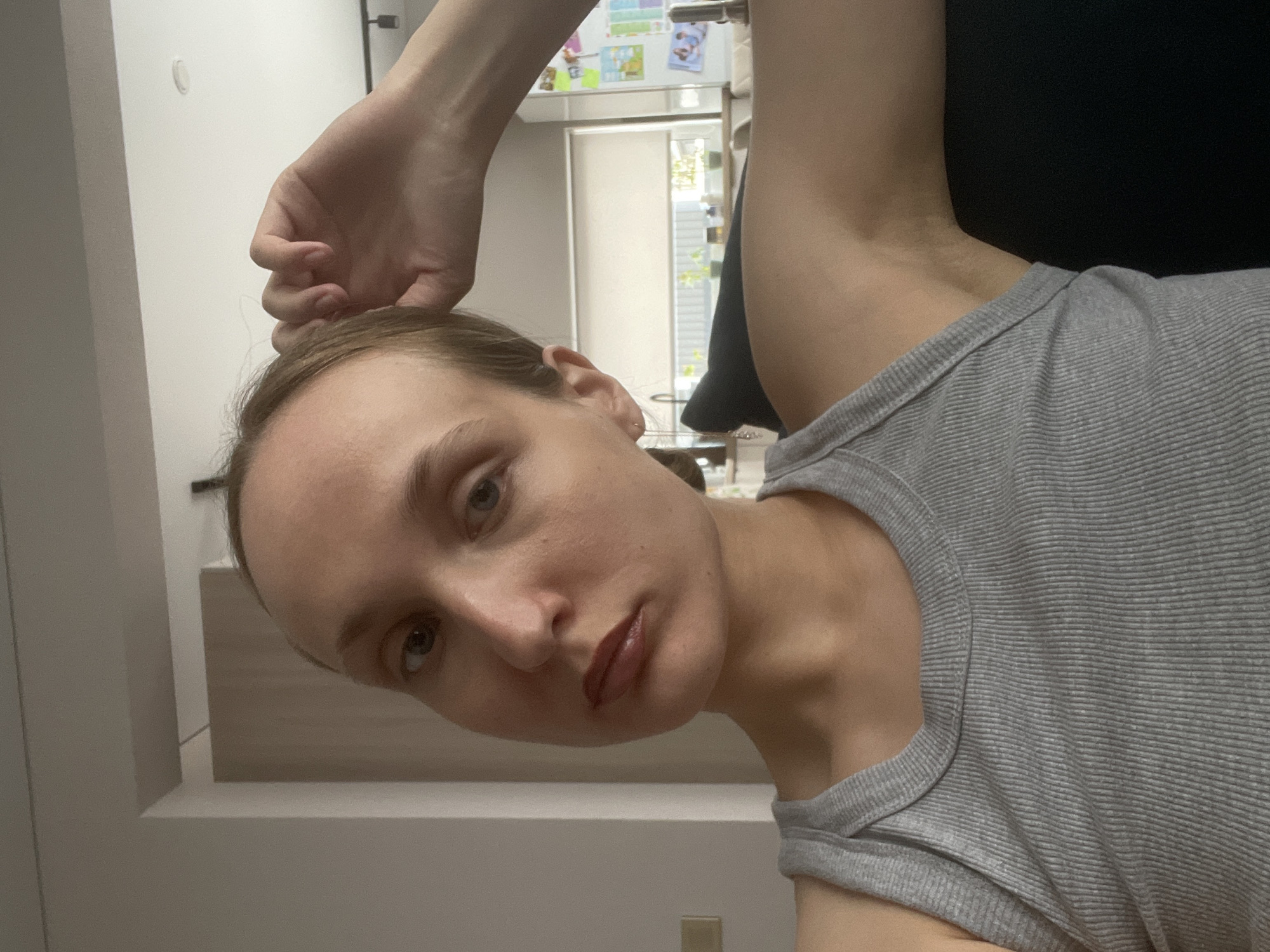A selfie shows a light-skinned person with brown hair and eyes. They wear a gray ribbed tank top. Their head is tilted downwards and to the left. A hand is raised above their head. The background includes a white wall, a refrigerator with papers and pictures attached, a window, and a dark surface. The location is potentially inside a dwelling in Sepych, Russia. No specific action or interaction is discernible.