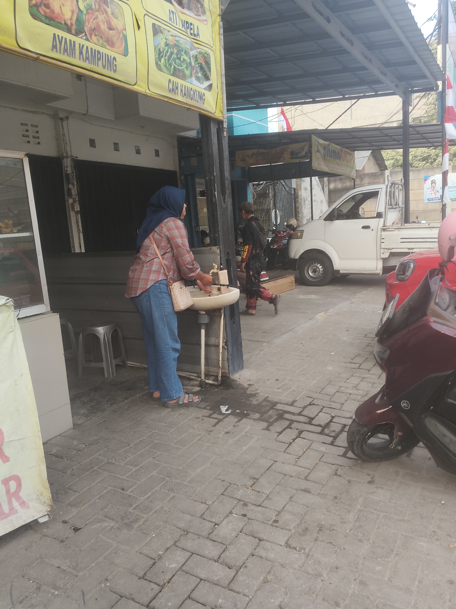 In Bandar Lampung, Indonesia, a woman wearing a blue hijab and a plaid long-sleeved shirt is washing her hands at a white sink on a cobblestone street. She is wearing blue jeans and gray sandals. A beige handbag is slung over her shoulder. Behind her, a young person in a black shirt and red pants walks away from the camera. In the background, a white pickup truck is parked next to a red motorcycle. A yellow banner with Indonesian text and images of food hangs above a building. The scene appears to be in front of a food stall or restaurant.