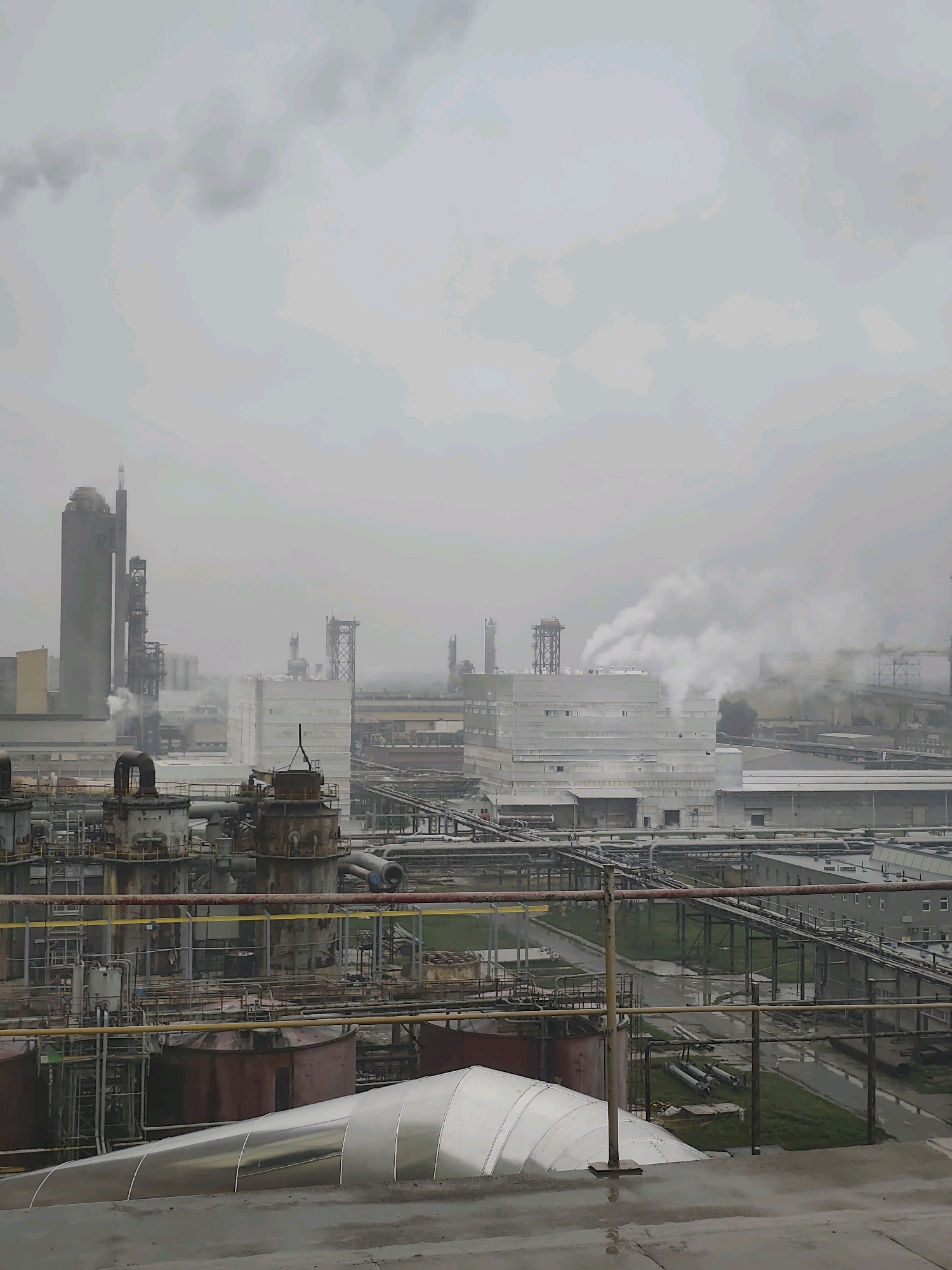 This image depicts an industrial complex, likely a chemical plant, under an overcast sky. The foreground shows a metallic, curved structure, possibly a pipe or tank, with a wet, concrete surface in front of it. In the mid-ground, a network of industrial infrastructure is visible, including large cylindrical tanks, numerous pipes, scaffolding, and elevated walkways. Several tall, industrial structures, including silos and processing towers, dominate the background. Plumes of white smoke or steam are emanating from some of these structures, indicating ongoing industrial processes. The overall atmosphere is one of industrial activity and a somewhat hazy, perhaps polluted, environment. The location is an industrial area of Police, Poland.