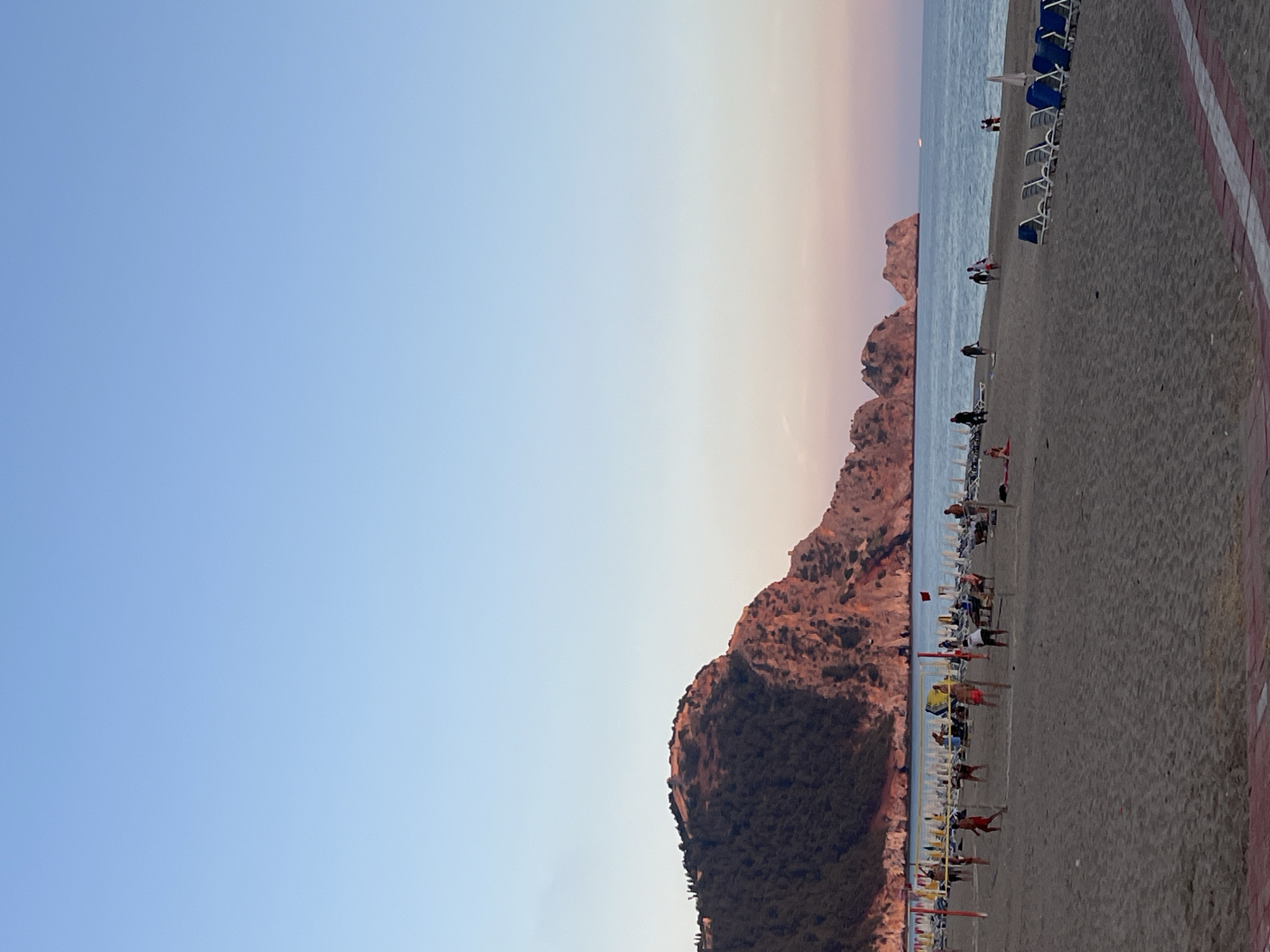 The image presents a panoramic view of a beach scene in Alanya, Turkey, likely during either sunrise or sunset, indicated by the sky's gradient from clear blue overhead to a soft pinkish-orange hue near the horizon.
To the left, a large, rocky mountain or hill, partially covered with sparse greenery, dominates the landscape, extending into the calm, bluish-green sea. Further out in the water, smaller rock formations are also visible.
The foreground and midground are filled with a wide, sandy beach. Numerous sun loungers, some with white frames and blue fabric, others entirely white, are neatly arranged in rows along the shoreline. A few beach umbrellas are also scattered amongst the loungers. People are visible across the beach as small figures; some appear to be walking, while others are standing or relaxing near the loungers. A few individuals are also seen in the distant water.
Along the very edge of the foreground, a paved walkway or promenade with distinctive red and white striped markings runs parallel to the beach. The overall setting is tranquil, capturing a peaceful moment at a popular coastal resort. There is no visible text in the image.