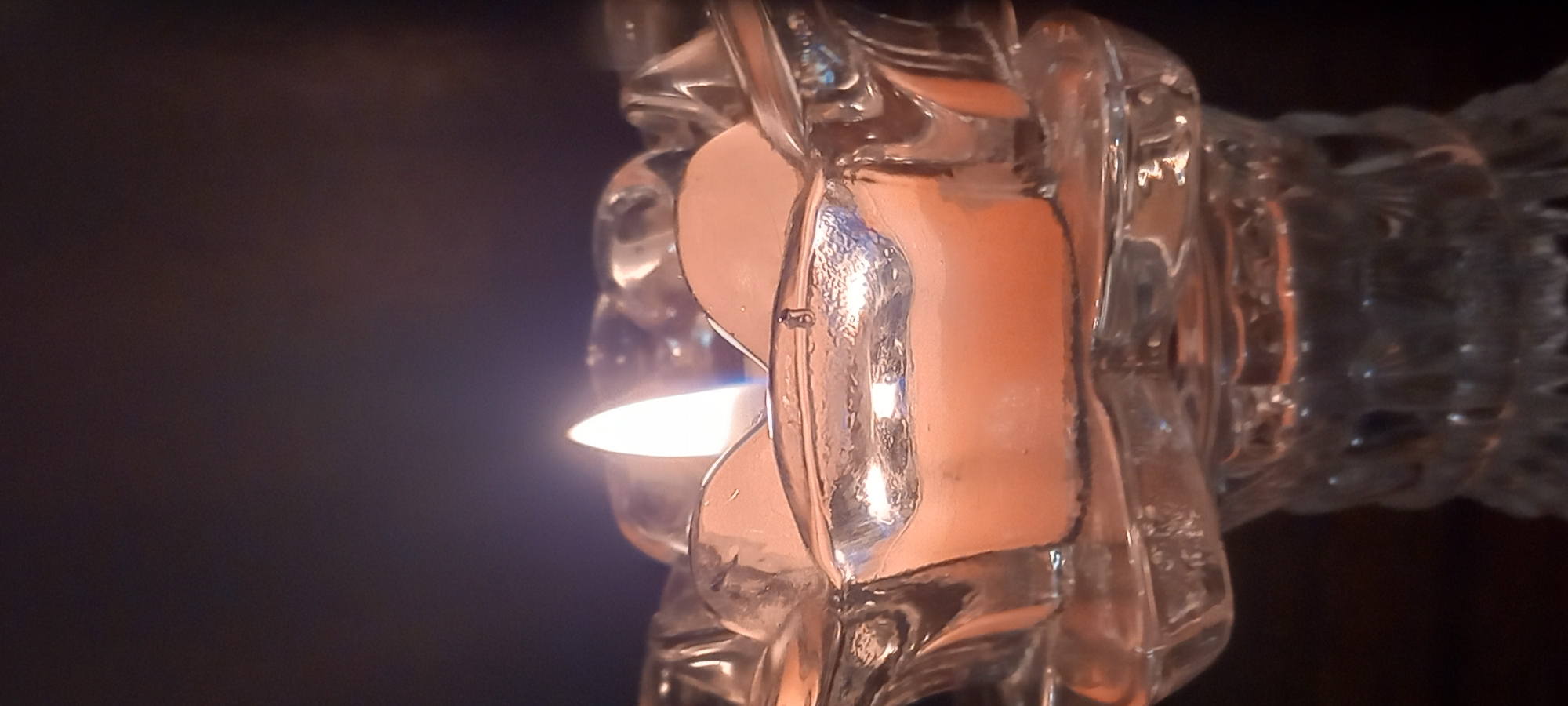 The image captures a close-up of a translucent, faceted object, likely made of glass or textured plastic, serving as a decorative piece or a candle holder. A small, lit candle or tea light is positioned behind or within the object, emitting a soft, warm, white and orange glow. The light from the candle refracts and diffuses through the multi-faceted surfaces of the object, casting a vibrant orange-amber hue across its inner and outer planes and creating intricate patterns of light and shadow.
The setting appears to be indoors, likely in a dimly lit room, as the background is dark and indistinct, emphasizing the illuminated object. The light source creates a peaceful and ambient atmosphere. The time of day is inferred to be evening or night, given the reliance on the candle's illumination. Faint, unidentifiable text or markings appear to be embossed or etched into the surface of the translucent object in several places. The image's location context is Kryvyi Rih, Ukraine. There are no people visible, and the primary activity is the burning of the candle.