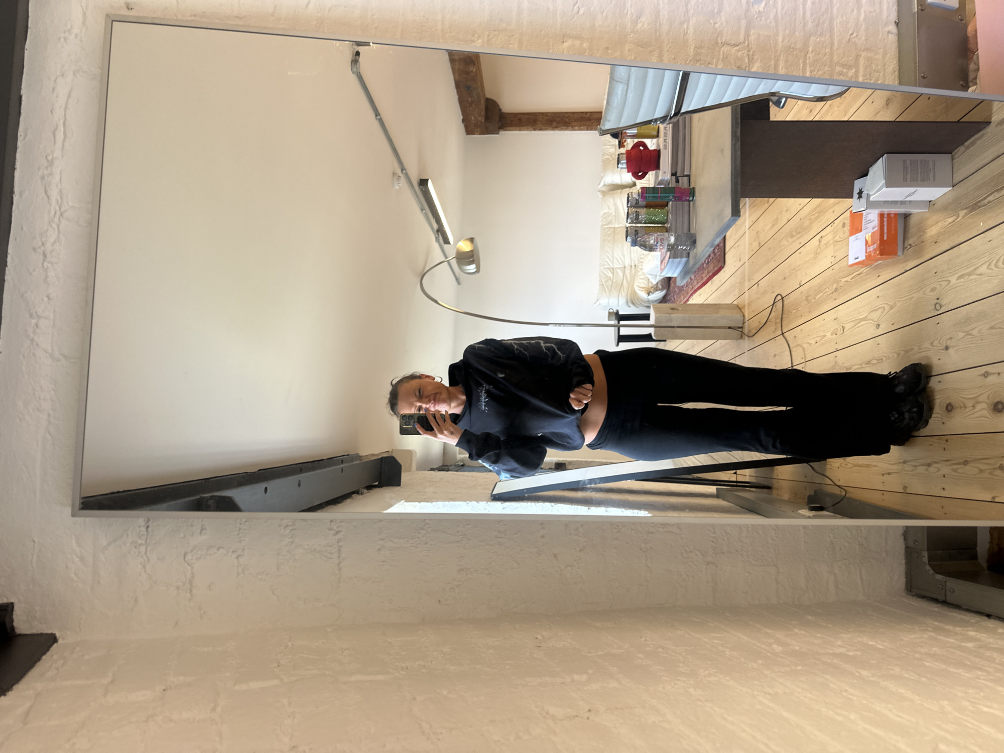 The image shows a full-body mirror selfie taken by a woman in an indoor setting. The scene is brightly lit, likely during the daytime.
**Subjects:** The main subject is a woman, visible in the mirror's reflection. She has dark hair pulled back and is wearing a black long-sleeved top, possibly with a subtle lightning bolt design, and black leggings or pants, complemented by dark shoes. She holds a smartphone to capture the photo, displaying a subtle, pleasant smile.
**Scene and Setting:** The room features light-colored wooden plank flooring and white-painted brick walls, giving it a rustic or industrial character. A large wooden beam is visible across the reflected ceiling. The large mirror itself has a thin, light-colored frame.
**Objects in Reflection:** The mirror reflects a living space. Visible objects include a bed or sofa with white bedding and cushions. A modern floor lamp with a curved metallic arm and a wall-mounted fluorescent light fixture illuminate the area. A dark wooden table or console holds various items: several clear bottles with colorful contents (one partially labeled "GLITTER"), a red decorative object, an Evian water bottle, and some books or magazines. On the floor, an orange box and a white box are visible, with "www.book-it.com" partially legible on the white box. A star-shaped ornament is placed beside the boxes. A small rug with a red pattern and a power cord are also seen on the wooden floor.
**Activity or Event:** The woman is casually taking a full-length self-portrait in the mirror.
**Notable Details:** The architectural elements like the exposed wooden beam, brick walls, and wooden floors suggest the location might be a renovated older building, consistent with the London, United Kingdom context. The atmosphere is casual and relaxed.