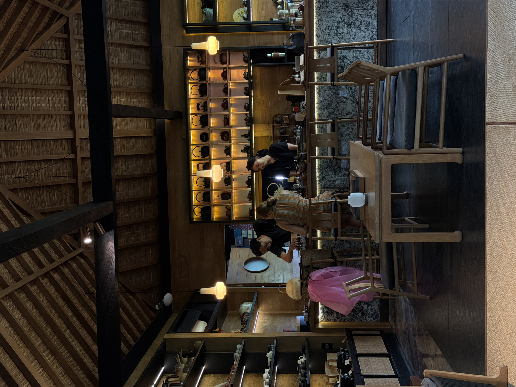 The scene depicts the interior of a tea house or cafe in Singakerta, Indonesia. Three individuals are present: one female customer and two staff members.
The interior features a high, peaked thatched roof with visible dark wooden support beams. Below the roof, dark horizontal wooden beams span the space. The walls are paneled with warm-toned wood. On the left, a tall, dark wooden shelving unit with integrated horizontal strip lighting displays various ceramic and tea-related items, including teapots, cups, and packaged goods.
Centrally located is a long service bar with a mottled grey-and-white patterned stone facade. Behind this bar, two staff members are visible. A male staff member, dressed in a dark shirt, leans forward over the counter, appearing to interact with the customer. A female staff member, also in a dark shirt, stands behind the counter to the right of the male, facing generally towards the customer area.
The female customer, wearing a light-colored, patterned garment, is seated on a wooden bar stool at the counter, facing the staff members. On the counter in front of the customer, various tea preparation items are visible. Directly in front of the counter, a light pink item, possibly a garment or bag, is draped over a wooden chair.
Behind the service bar, a large wooden shelving unit with individual illuminated compartments displays numerous dark-colored teapots and smaller tea ware items. To the right of this unit, another shelving area shows more items.
In the foreground, a light-colored wooden table with two wooden chairs, each featuring dark seat cushions, is positioned. A small white ceramic object rests on this table. The floor is dark-colored, possibly slate or dark tile, with a lighter-colored, textured mat or rug extending into the immediate foreground. Several pendant lights with opaque, light-colored shades hang from the ceiling, illuminating the bar and seating areas.