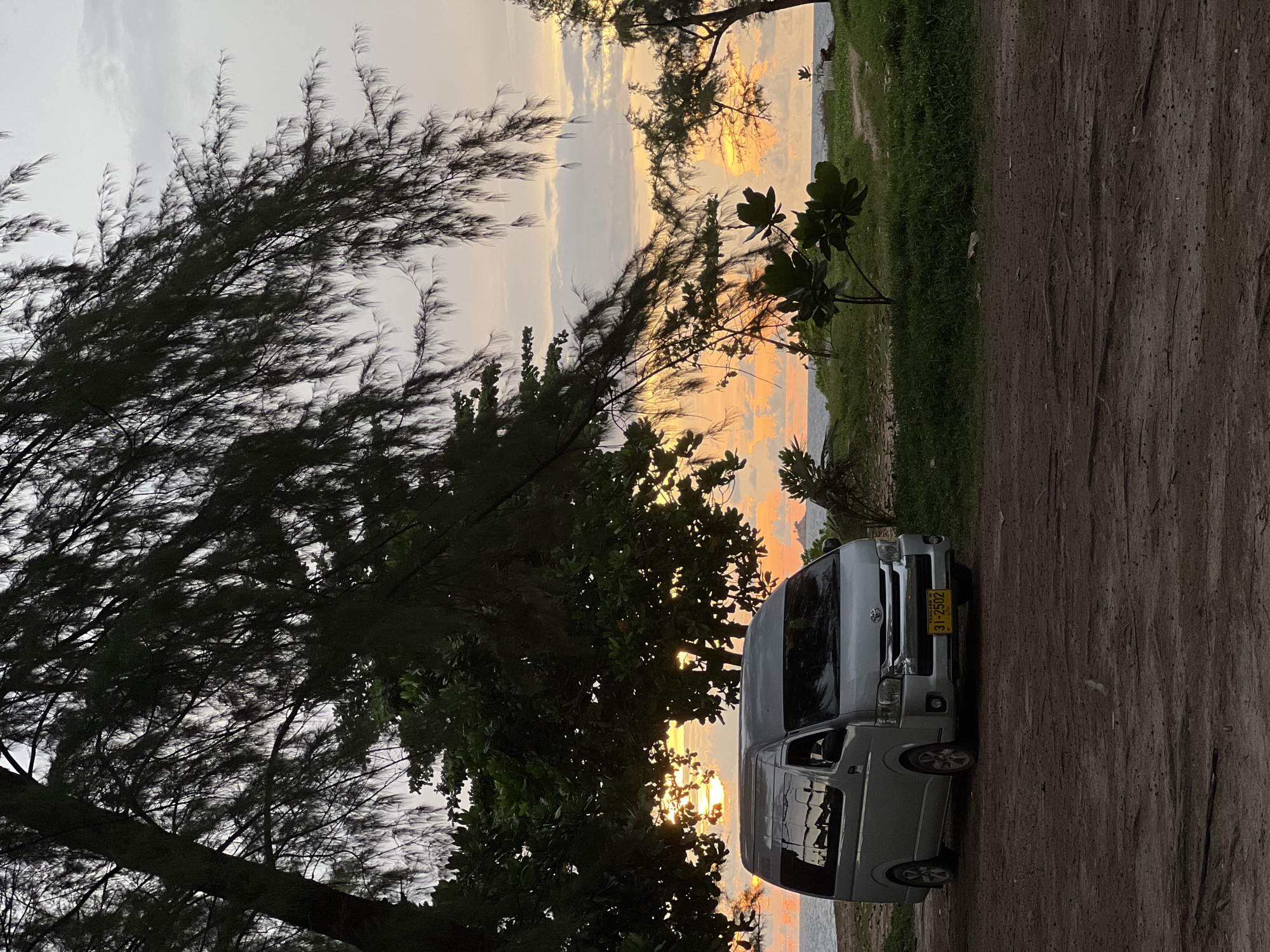 A silver minivan is parked on a dirt lot near a beach during sunset. The van is facing the ocean, and its headlights are on. Trees and foliage surround the van, with the ocean visible in the background. The sky is filled with orange and yellow hues, indicative of a sunset. The license plate on the van reads "31-2502 THAILAND". There are no people visible in the image. The location is unknown.