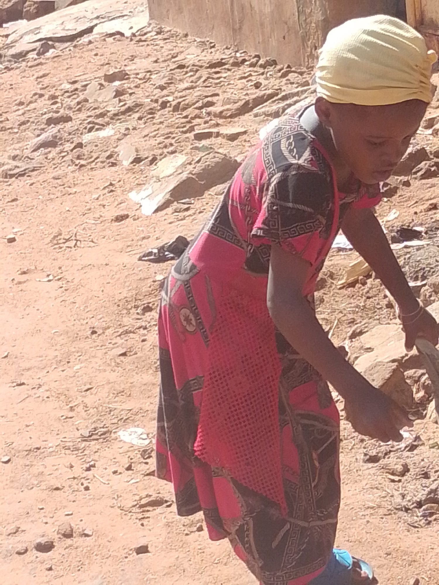 A young person wearing a yellow headscarf and a pink and black patterned dress is shown from the side, bending over to interact with the ground. The ground is a dusty, sandy terrain with scattered rocks and debris, suggesting an outdoor, possibly rural or undeveloped, environment. The person's right arm is extended, holding a wooden stick or tool, which they are using to move or examine something on the ground. Their head is bowed, indicating focus on the task at hand. The lighting suggests it is daytime, with bright sunlight casting shadows. The person's attire is simple, and their posture implies they are engaged in some form of labor or activity related to the ground. No text is visible in the image.