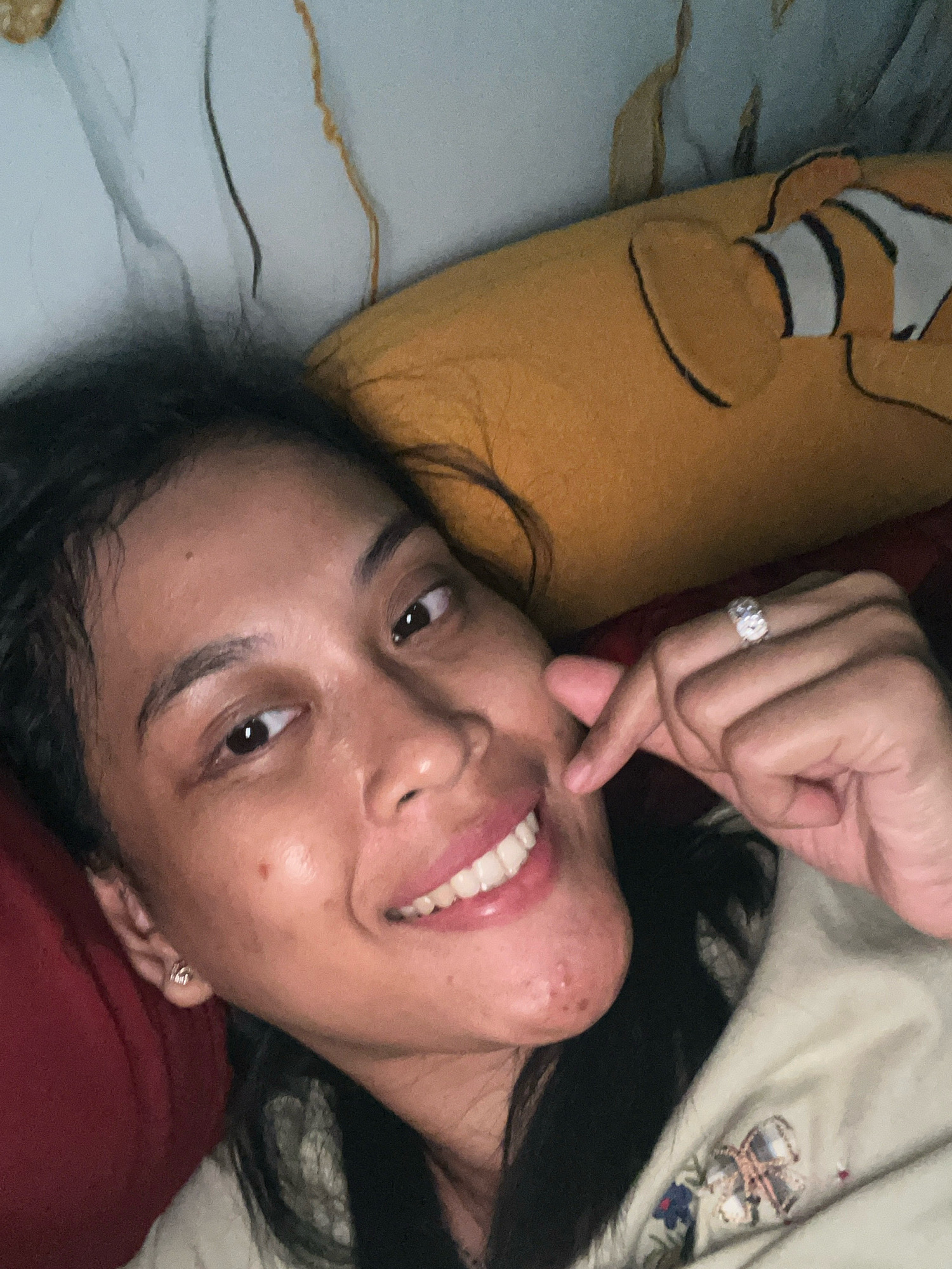 A woman is lying down, taking a selfie. She is smiling and touching her cheek with her right index finger, which is adorned with a ring. Her left earlobe has a small earring. She is wearing a light-colored top with a pattern. Behind her, a yellow pillow with a clownfish design is visible, along with what appears to be bedding. The background shows a textured surface, possibly a wall or fabric. There are no elements in the image that suggest a specific location in Kecamatan Pati, Indonesia.