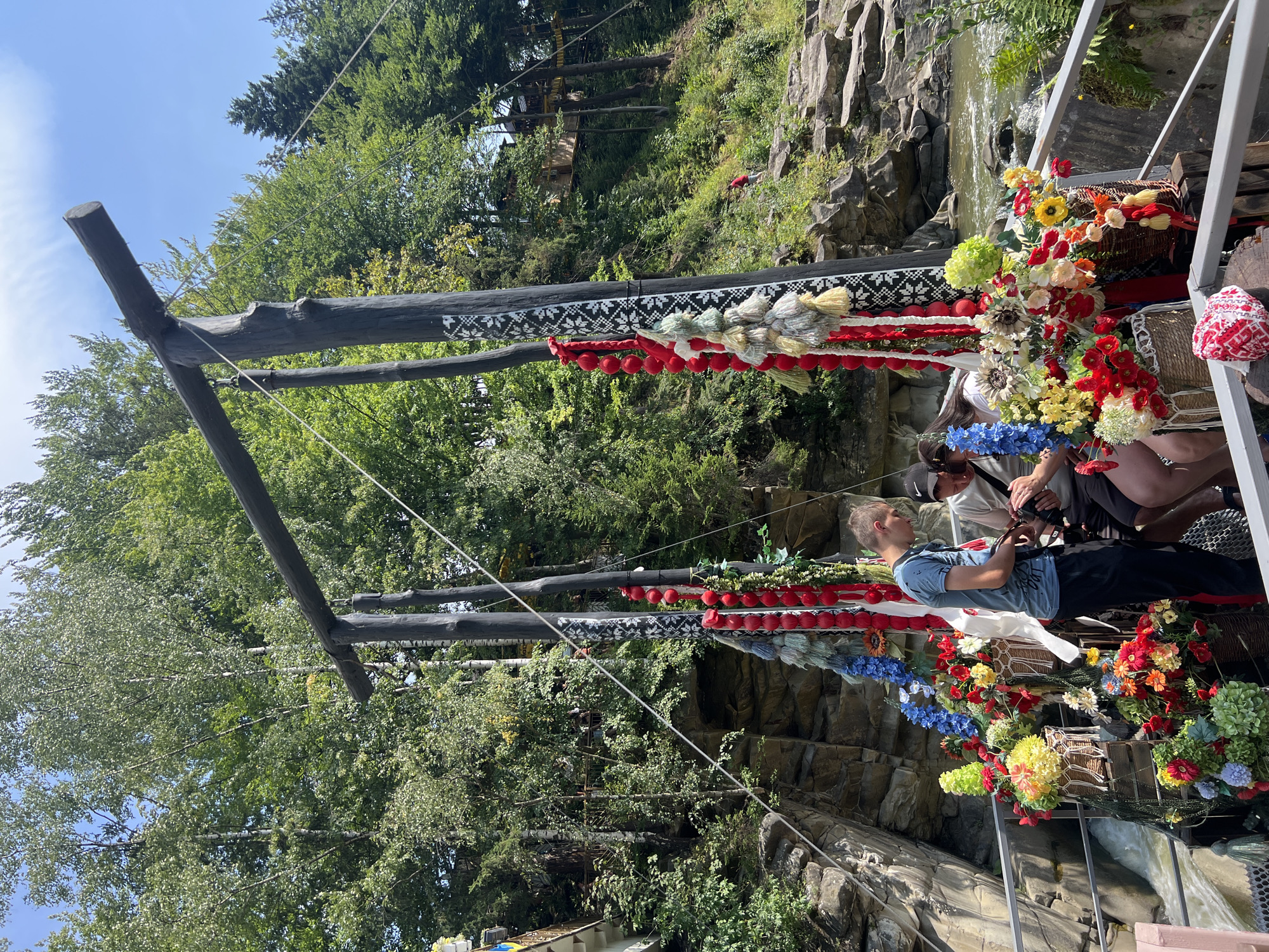 This image depicts a scene in Yaremche, Ukraine, focusing on a wooden structure adorned with vibrant floral arrangements and traditional decorative elements, possibly related to a local festival or cultural event. The structure, resembling a large swing or a decorative archway, is made of dark, rough-hewn logs. Hanging from the crossbar are strings of red spheres and tassels in various muted colors, interspersed with black and white woven patterns.
Below the main structure, on what appears to be a platform or bridge, are several individuals. A young man in a blue t-shirt and black pants is standing and looking down. To his right, two people are partially visible, one wearing a black baseball cap and the other holding a camera, suggesting they might be tourists or participants. The platform is heavily decorated with numerous baskets overflowing with artificial flowers in shades of red, yellow, blue, green, and white. The flowers are arranged in a lush, overflowing manner, creating a colorful and festive display.
The background reveals a natural landscape characteristic of Yaremche, with a fast-flowing river or stream cutting through a rocky gorge. Steep, forested hills rise on either side of the river, densely covered with green trees. The sky is a clear blue with scattered white clouds. The overall impression is one of a lively, decorated outdoor setting within a picturesque natural environment.