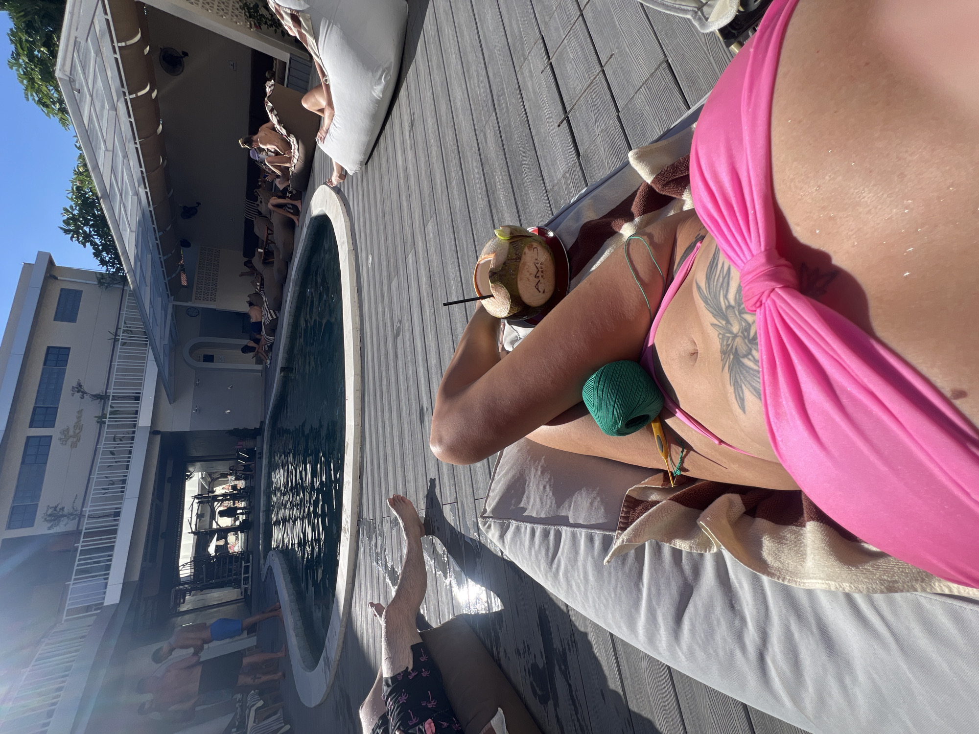 The image depicts a scene next to a pool in what appears to be a resort or recreational area. A person is reclining on a white lounge chair, wearing a pink bikini and a tattoo. They are holding a coconut drink with a straw, next to a green ball of yarn and a crochet hook.
Next to the lounge chair, another person's legs and feet are visible, indicating they may be on another lounge chair. The pool is bordered by a wooden deck and a row of bean bag-like chairs. Several people are in or around the pool, with a building visible in the background and a sky with sunlight.
Given the location information, this scene is likely in North Kuta, Indonesia.