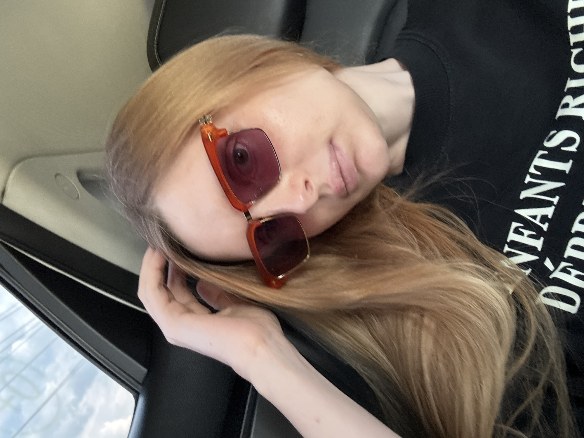 A fair-skinned woman with blonde hair and sunglasses with orange frames is looking forward, with her left hand on her head, while sitting in a car. She is wearing a black t-shirt with white text that reads "ENFANTS RICHES DEPRIME." The background shows the interior of the car and clouds through the window. The image is taken from a medium close-up angle.