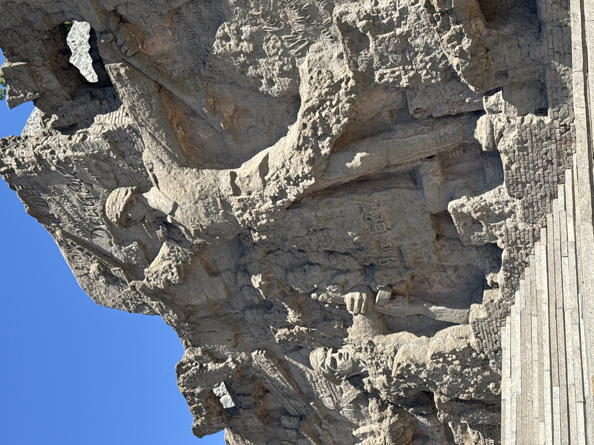 The image shows a close-up, low-angle view of a monumental relief sculpture on a rock face against a bright blue sky. The sculpture depicts several figures in military attire, carved from the rock itself. The style is realistic and imposing, with deep carving creating strong shadows. In the foreground, a wide set of stone stairs leads up to the base of the monument, which is constructed of rough-hewn stone and brickwork. The sculpture features a prominent central figure, appearing to be a male soldier with a helmet, his body angled upwards. To the left, another figure, possibly a woman or another soldier, is depicted, also carved from the rock. The rock face is textured and appears weathered, with visible cracks and indentations. There are inscriptions carved into the rock face in a script that appears to be Cyrillic. This monument is part of the Mamayev Kurgan memorial complex in Volgograd, Russia, commemorating the Battle of Stalingrad.