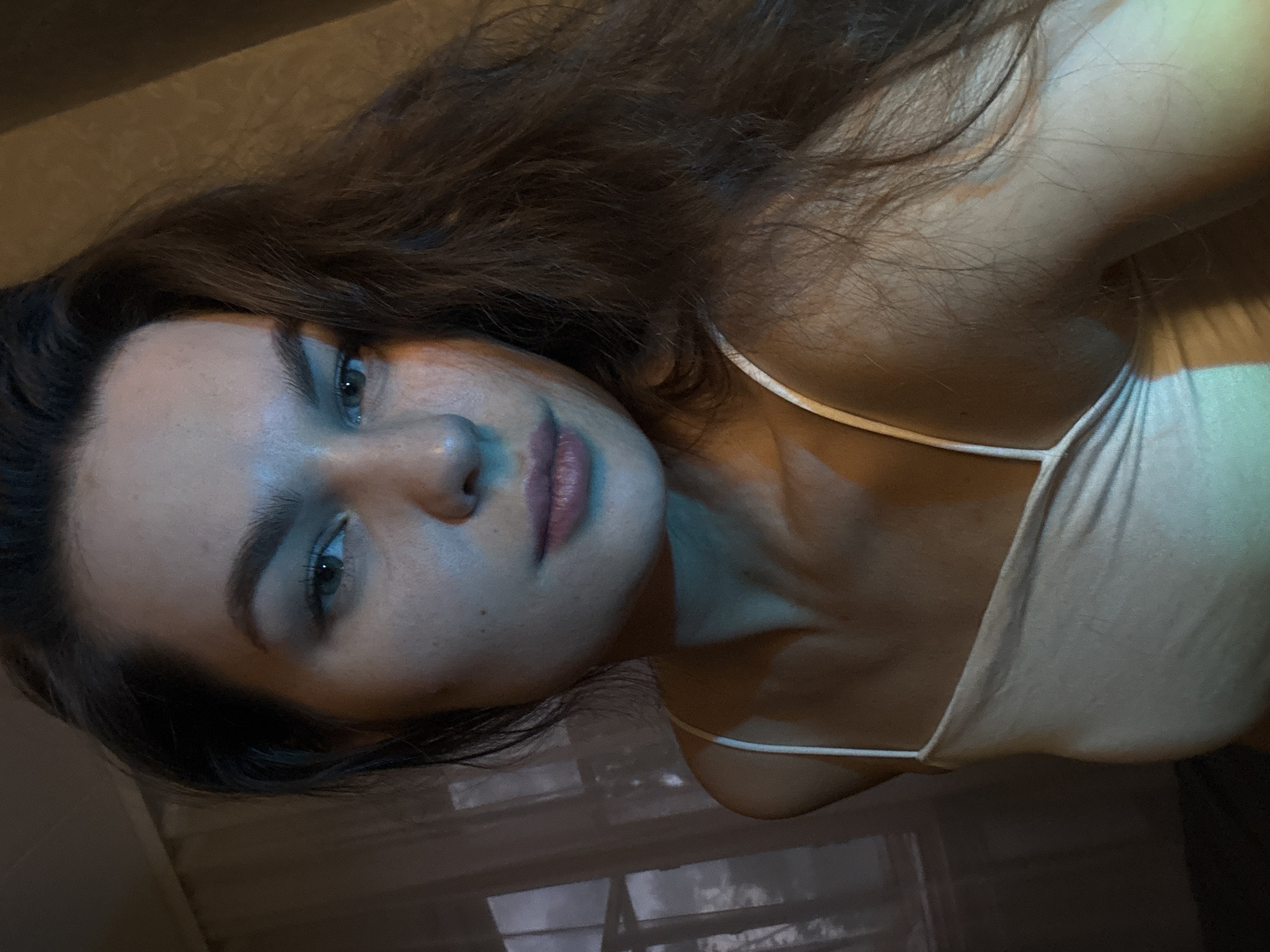 A young woman with long, dark brown hair is pictured in a close-up, selfie-style shot. She is wearing a white tank top with thin spaghetti straps. The lighting in the image is dramatic, with blue and green hues casting shadows and highlights across her face and upper body. Her skin appears to have some light freckles. She is looking directly at the camera. The background is dimly lit and appears to be an indoor setting, possibly a bedroom, with a window and sheer curtains visible to the left. The overall impression is one of soft, ambient lighting with a slightly moody atmosphere. The location is not specified in the image itself.