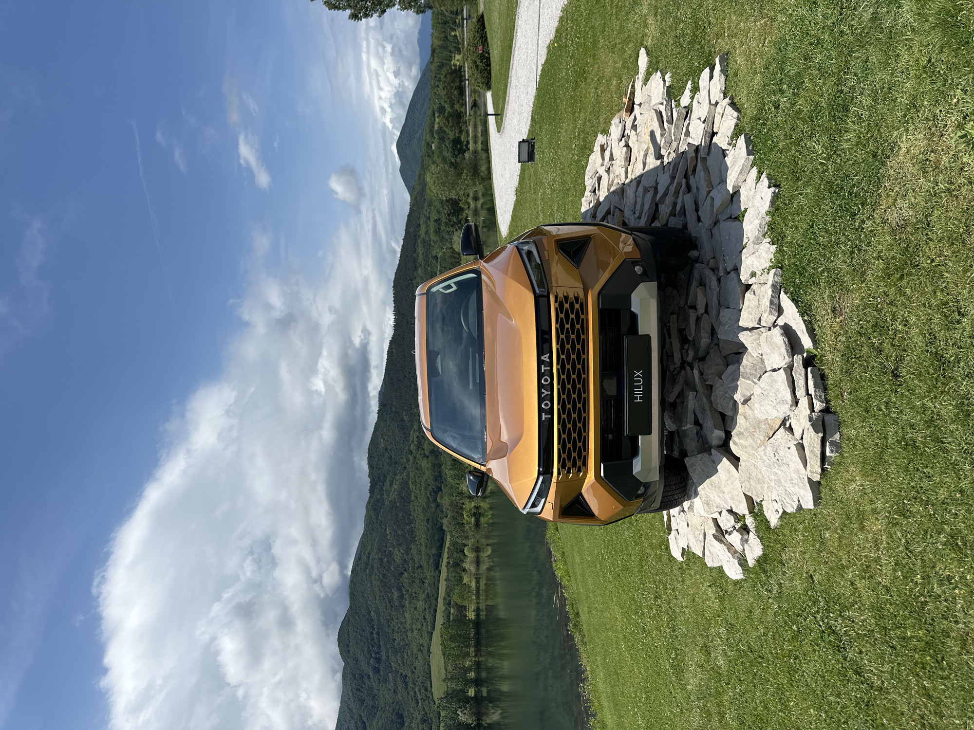 A golden-orange Toyota Hilux pickup truck is prominently displayed outdoors in a scenic, mountainous landscape in Razliv, Bulgaria. The truck is positioned centrally in the foreground, facing slightly to the left, on a circular platform constructed from irregularly shaped, light-colored rocks. Lush green grass surrounds the rock platform and extends into the foreground and mid-ground.
In the mid-ground, a calm body of water, likely a lake or river, stretches horizontally, reflecting the blue sky and distant mountains. The background features impressive mountains, densely covered with green trees, rising under a clear blue sky adorned with scattered white clouds. A bright white gravel path curves into the distance on the right side of the image.
The scene appears to be set during bright daylight, possibly late morning or early afternoon, indicated by the strong sunlight and distinct shadows cast by the rocks and the truck. The weather is clear and sunny. No people are visible, suggesting the truck is on display or being photographed for promotional purposes.
Visible text on the front of the truck includes "TOYOTA" in large, capitalized letters across the grille, and "HILUX" on a black plate positioned below the grille. The overall setting evokes a sense of rugged natural beauty, highlighting the vehicle's design against a picturesque outdoor backdrop.