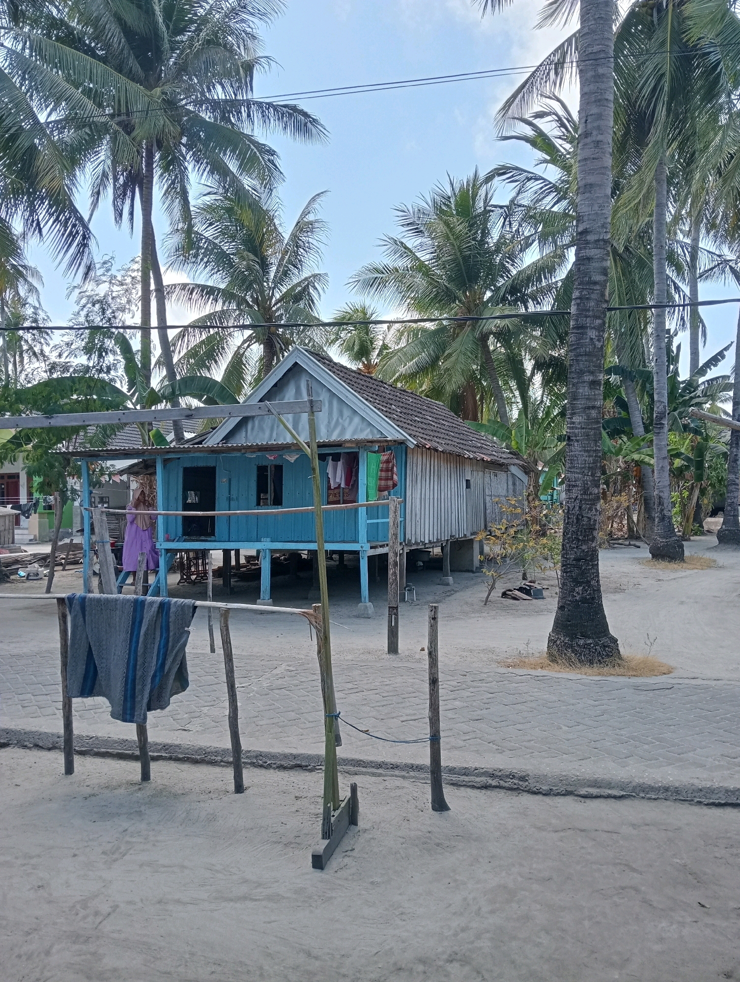 The image depicts a scene in Pagerungan Kecil, Indonesia, featuring a small, stilted wooden house with a blue-painted lower section and a tin roof. The house is surrounded by lush palm trees and other tropical vegetation. In the foreground, a clothesline holds a gray and blue striped blanket or rug. A person wearing a light purple garment is visible near the entrance of the house. The ground is sandy, with a paved walkway visible to the right of the house. The sky is clear and blue. The overall impression is that of a humble dwelling in a tropical coastal village.
