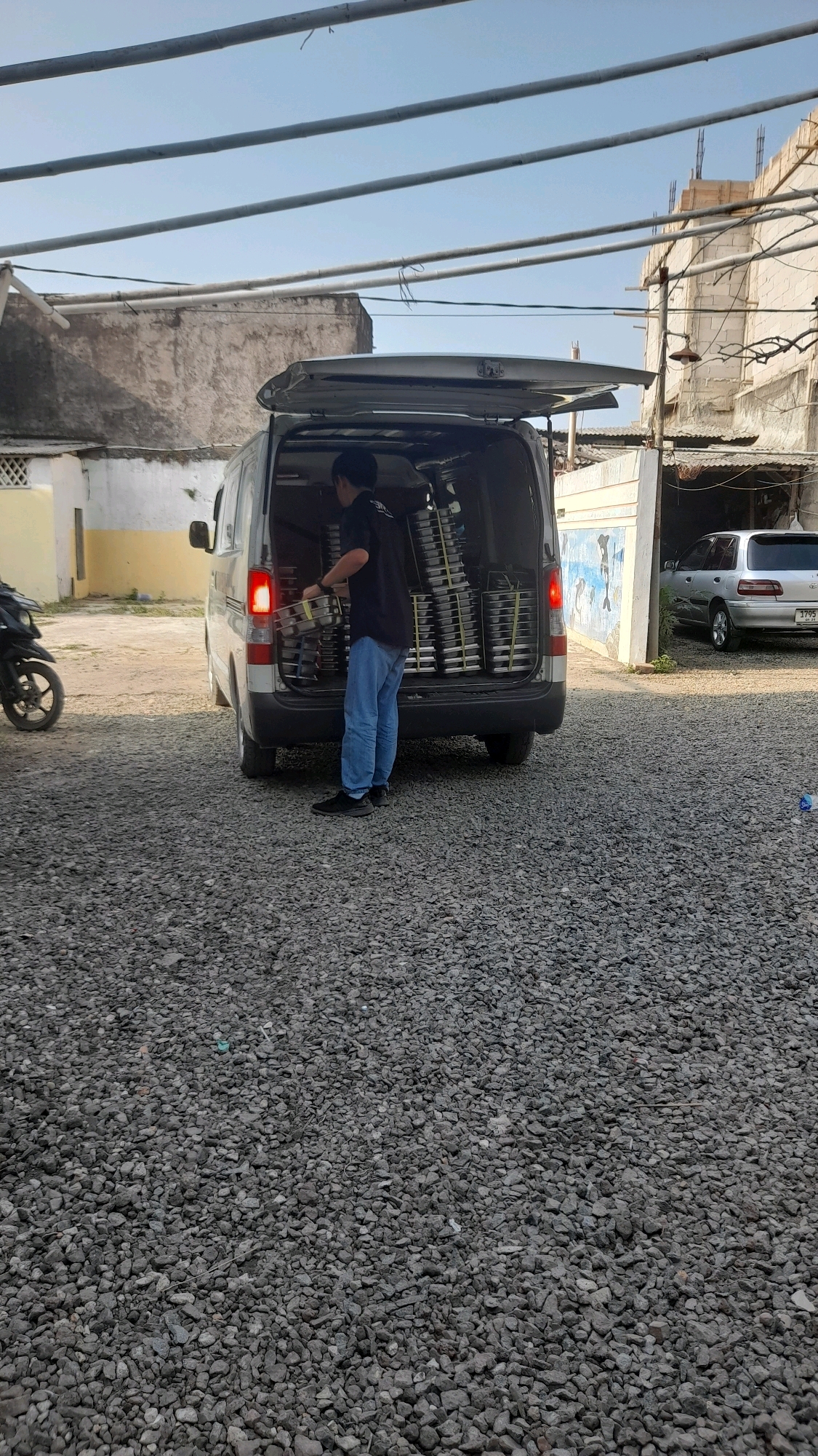 A person is standing next to the open back of a van and appears to be unloading stacked trays. The van is silver and has its rear doors open. The person is wearing a black shirt, blue jeans, and dark shoes. The background shows a gravel lot, a building with a mural on its side, and another parked car. The scene is outdoors during the day, with a clear sky. The location is in Selaawi, Indonesia.
