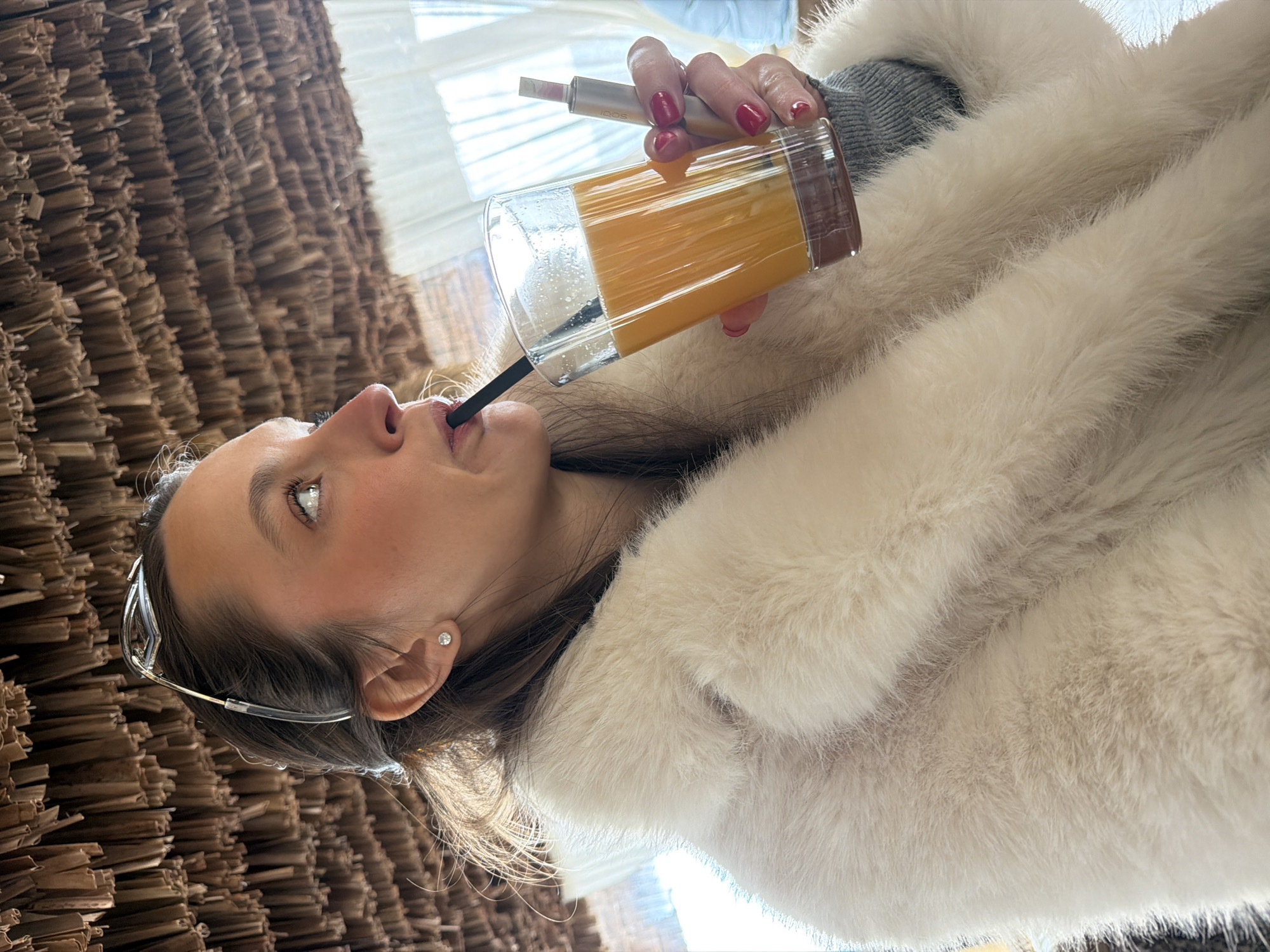 A woman in a white fur coat is drinking a yellow liquid through a black straw from a clear glass. She holds an electronic cigarette in her other hand. She is wearing eyeglasses and an earring. The photo appears to have been taken indoors, with a brown woven wall covering visible on the left side of the frame, and a window in the background. The location is unknown, although the prompt suggests it may be Petrovac centar, Montenegro.