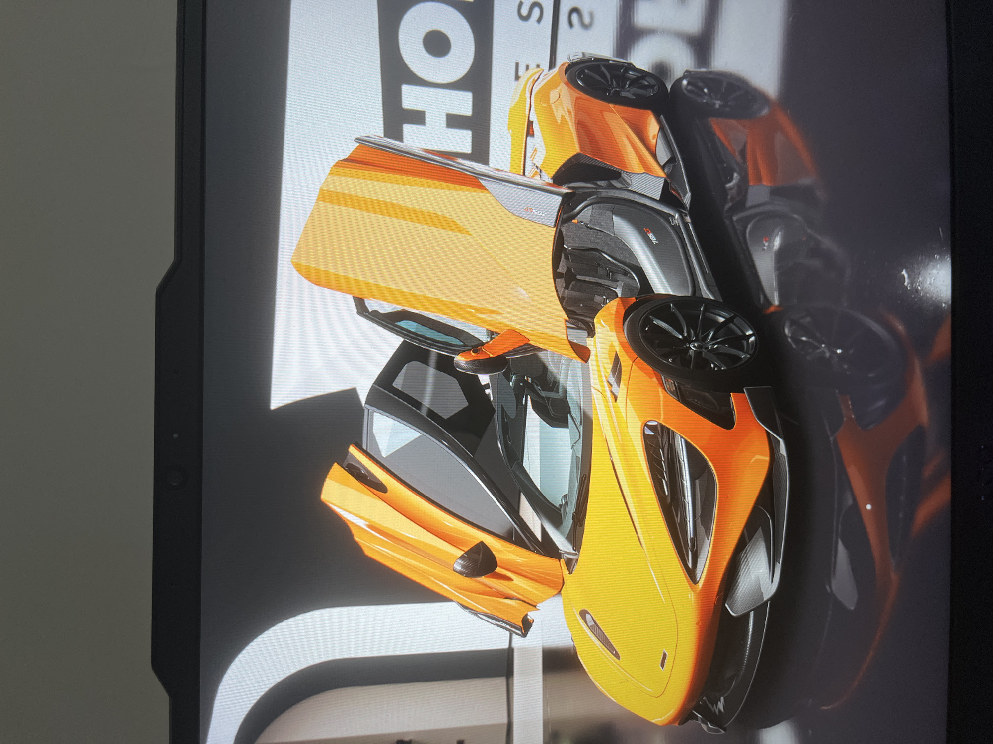 An orange McLaren 765LT Spider is on display. The doors are open, revealing the interior. The background features the letters "HO" and "S 2" in large, bold print. A reflection of the car is visible on the dark surface behind it. The location is in the city of Unknown, Unknown.