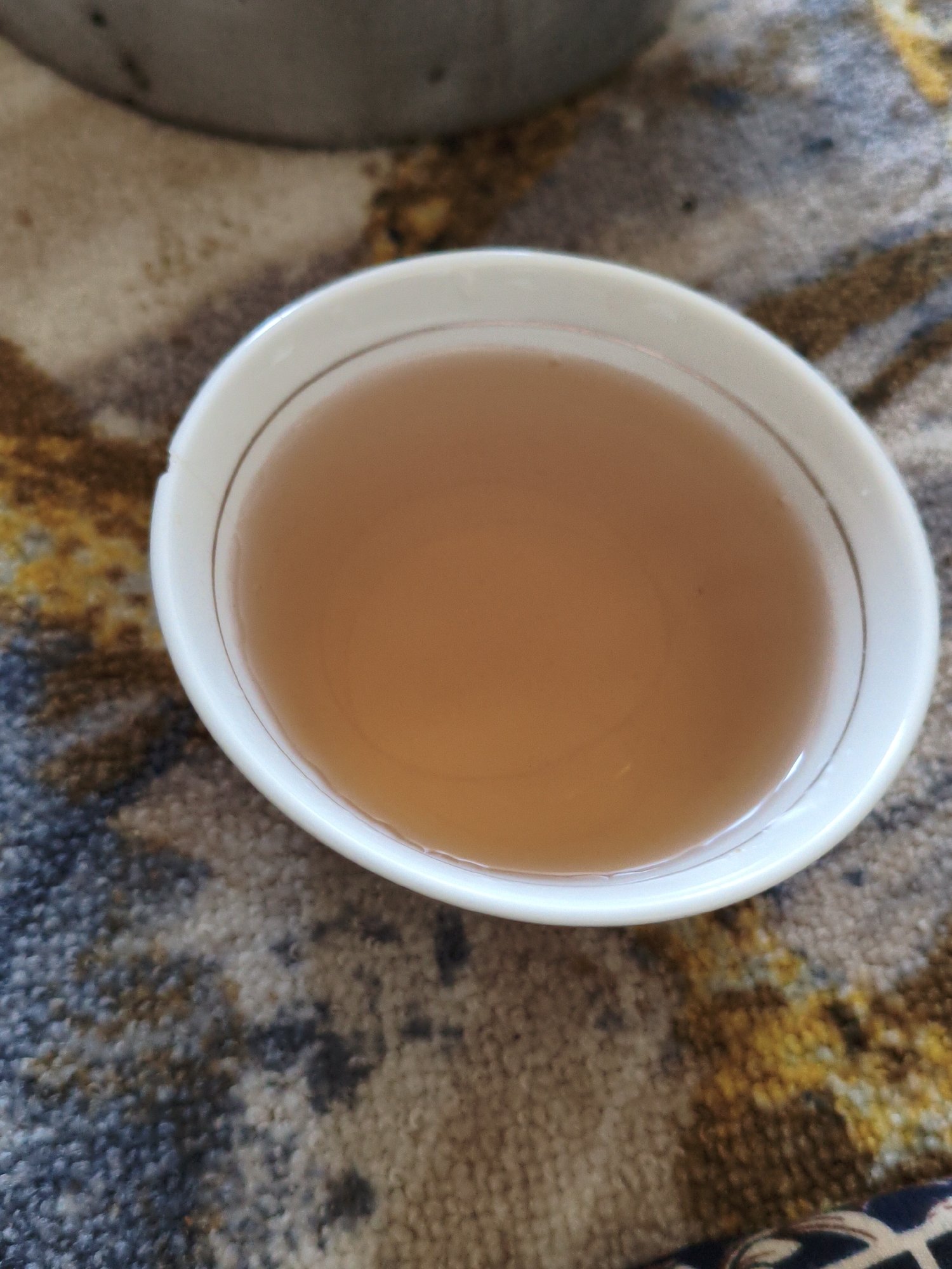 A white teacup with a thin gold rim filled with amber-colored liquid sits on a textured, patterned rug. The rug has a mottled design with shades of brown, blue, and yellow. In the background, out of focus, a portion of a grey metal object is visible, possibly a pot or container. The scene is indoors, with the lighting suggesting a natural, ambient light source. The image is a close-up shot, focusing on the teacup and its contents. The overall impression is one of a still life, with an emphasis on the warm tones of the liquid and the rug. There is no indication of the city of Механизатор, Tajikistan, or any specific location within it.