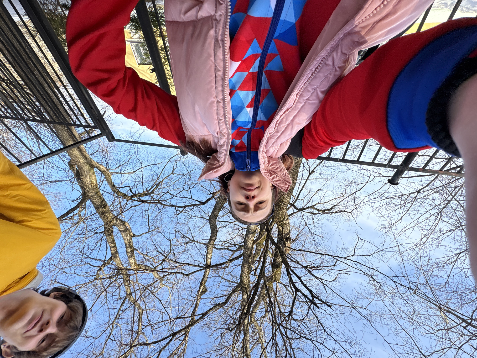 From a low angle beneath a tree, two individuals, one clad in a yellow jacket, the other in a red, pink, and blue jacket, are visible against the backdrop of bare branches reaching towards a light blue sky. A dark metal structure is partially visible in the upper left. The location is possibly Berezniki, Russia.