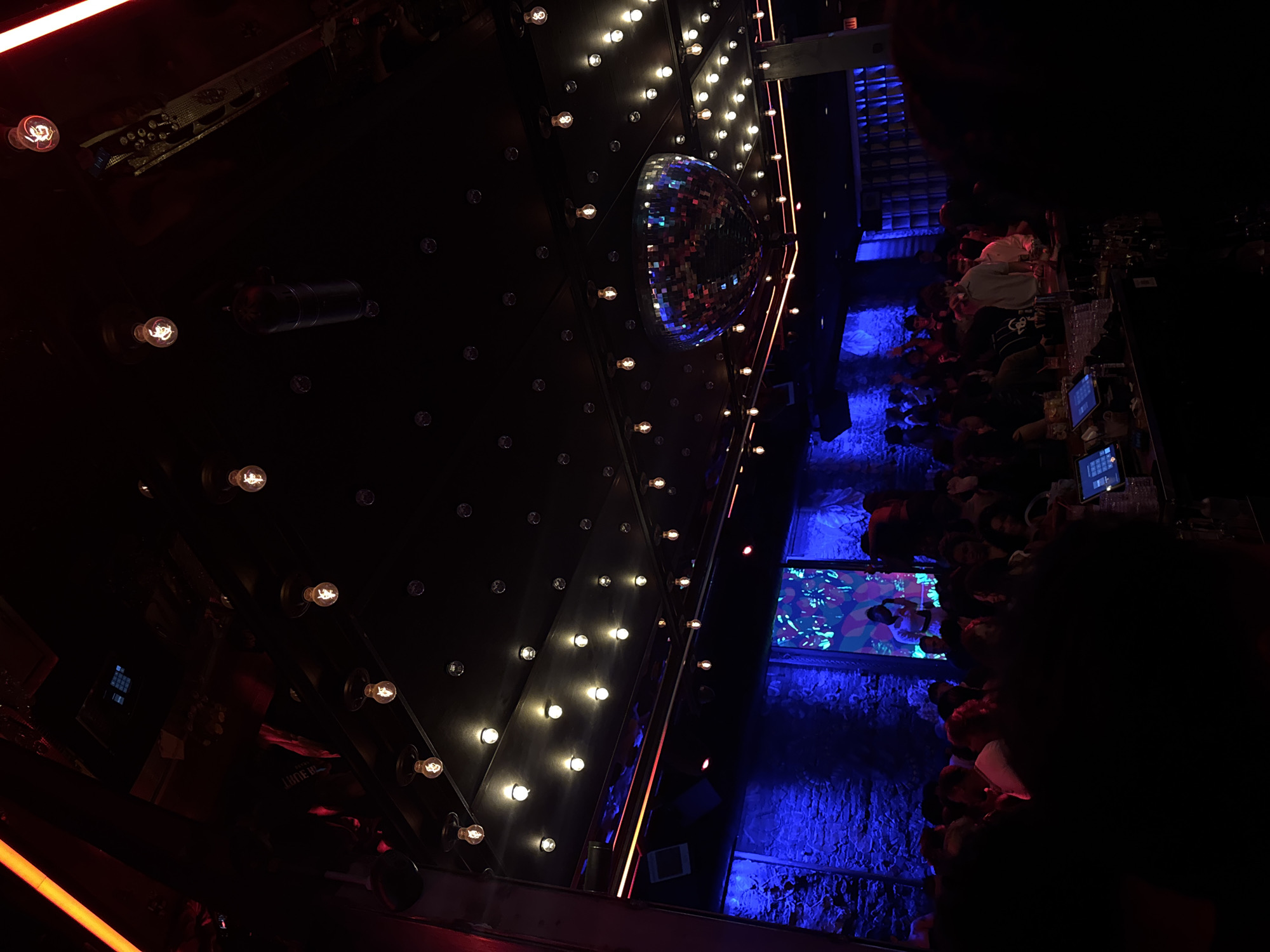 The image depicts a dimly lit interior, likely a bar or nightclub. Dominating the scene is a large, dark wall adorned with numerous light bulbs arranged in a geometric pattern. A disco ball hangs prominently, reflecting light onto the walls. Below, patrons are crowded around a bar, with several people visible and others obscured by darkness. The lighting is primarily blue, with some red and orange accents. The scene is likely located in a city setting, possibly Chicago, USA.