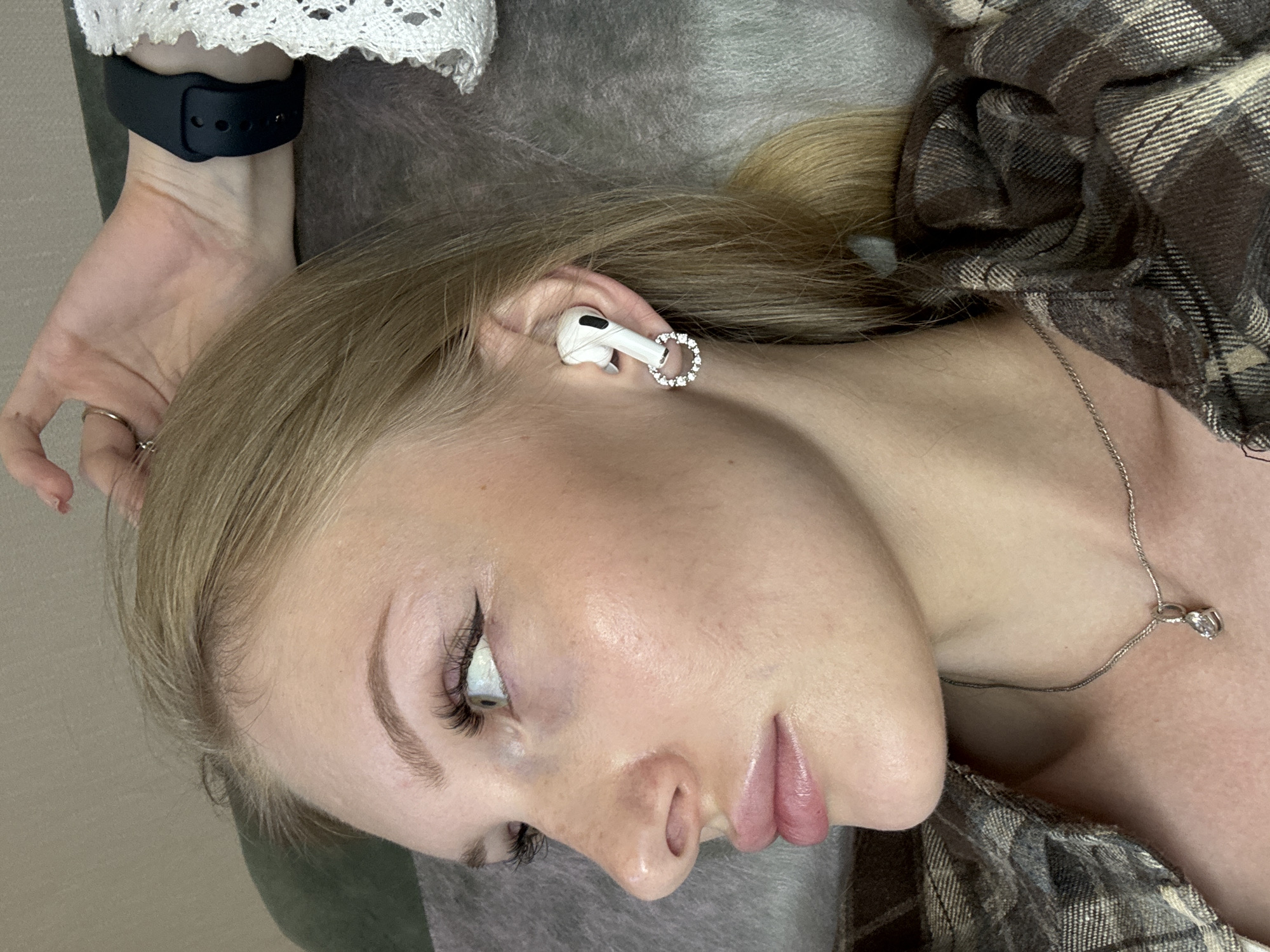 A young Caucasian woman with blonde hair is shown in a profile view, looking to the left. She is wearing a plaid shirt and a silver necklace with a pendant. Her left ear is adorned with a white earbud and a circular earring with small, clear stones. A dark smartwatch is visible on her right wrist. Her left hand is raised, touching her hair, and she is wearing a ring. The background is out of focus, appearing to be a textured surface, possibly a sofa or blanket. The image is cropped to focus on the woman's face and upper body. The specific location in Perm, Russia, cannot be determined from the visual information provided.