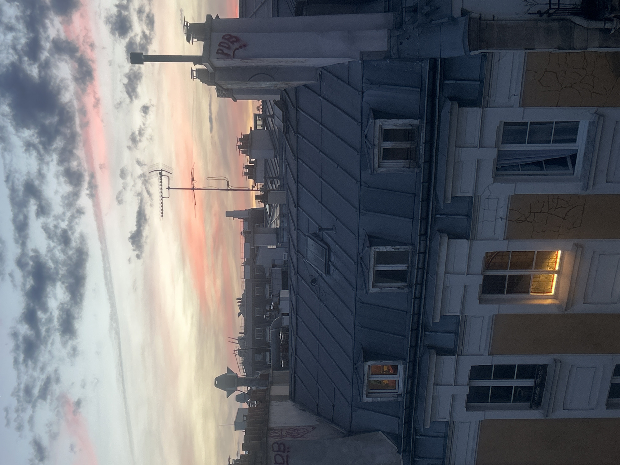 The image depicts an urban rooftop landscape under a colorful sky at either dawn or dusk. The sky, stretching across the left and top, features soft streaks of pink, orange, and white clouds blending into darker grey patterns.
In the foreground and mid-ground, a collection of multi-story buildings with characteristic dark grey zinc roofs and dormer windows dominates the scene. Numerous chimneys, vents, and television antennas, including a prominent Yagi-style antenna, punctuate the rooftops.
On the right side, the facade of a building is visible, featuring multiple windows with light-colored, possibly cracked, walls and decorative frames. One window on the middle-right emits a warm yellow light, indicating an illuminated interior. Another window just below it is slightly ajar.
Red graffiti spelling "PDB" is discernible on a tall, light-colored chimney or vent stack. The overall atmosphere is tranquil, capturing a quiet moment in a city environment, likely a European one given the architectural style. No people or overt activities are visible, emphasizing the stillness of the moment.