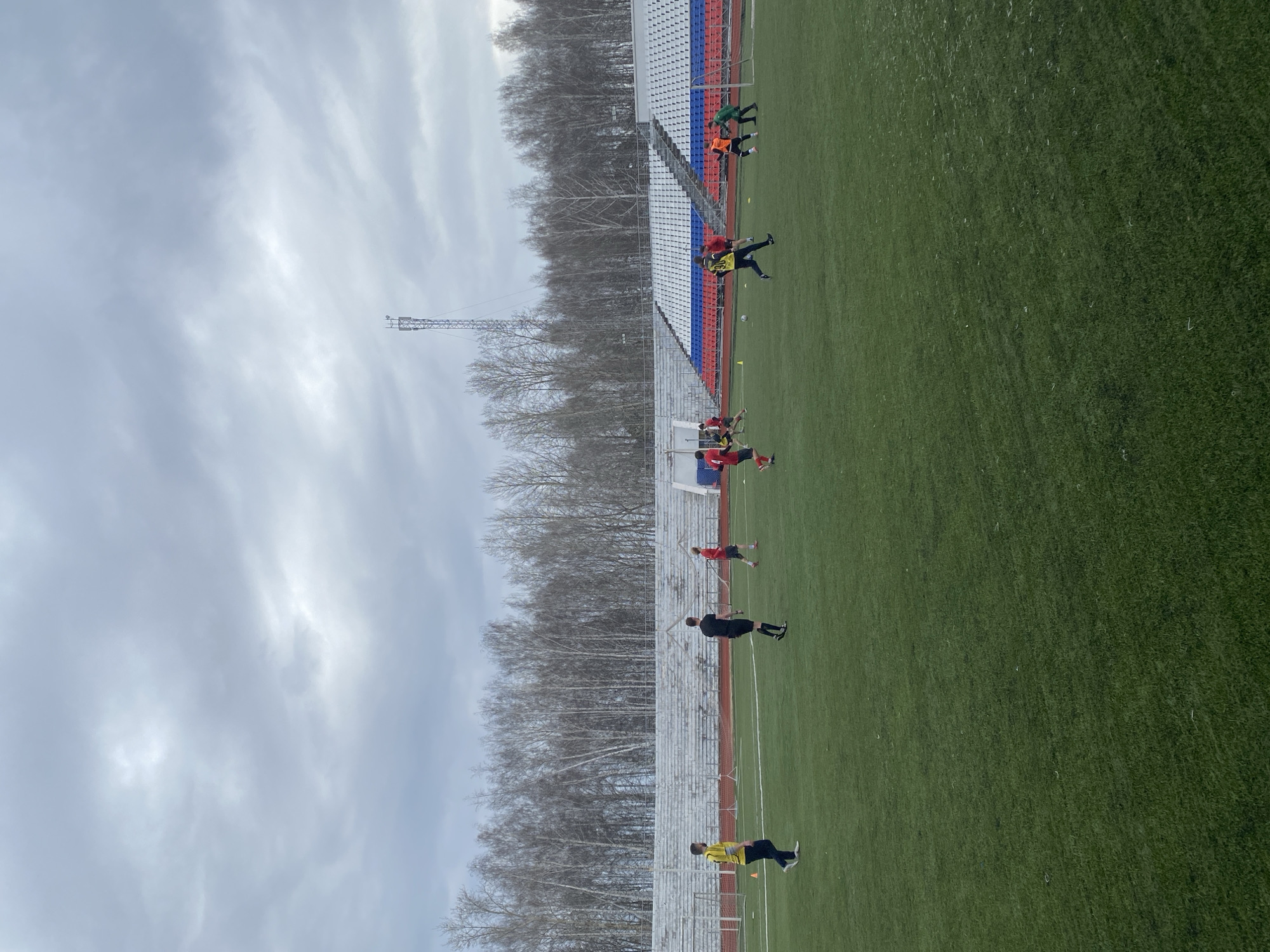 The image shows a soccer field in Chaikovsky, Russia, with a row of bleachers to the left and a row of bare trees behind them. A metal tower can be seen near the bleachers. There are several people on the soccer field wearing athletic clothing, some in yellow, some in red, some in black, some in orange, and some in green. They appear to be practicing or warming up on the field. The sky is overcast.
