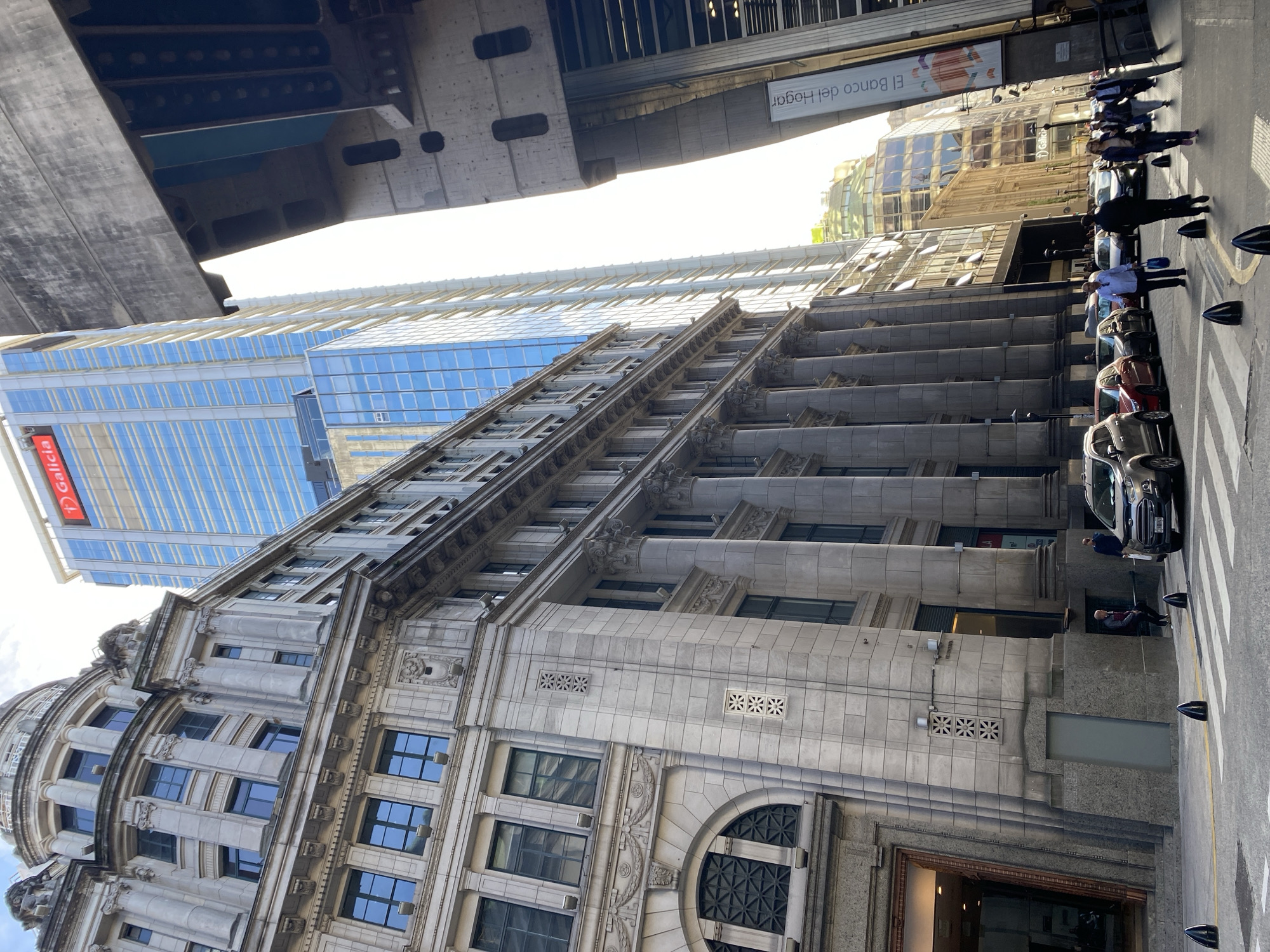 A ground-level shot taken in Buenos Aires, Argentina, features a tall, ornate stone building with arched windows and detailed architectural elements, juxtaposed with a modern glass skyscraper visible behind it, with a “Galicia” bank sign. Above the building, there is a concrete structure, potentially a bridge or elevated road. On the street level, multiple parked cars and pedestrians are observed, some walking on the sidewalk. A Banco del Hogar advertisement is also visible on the side of another building. The image seems to be taken from a low angle, emphasizing the height of the buildings.