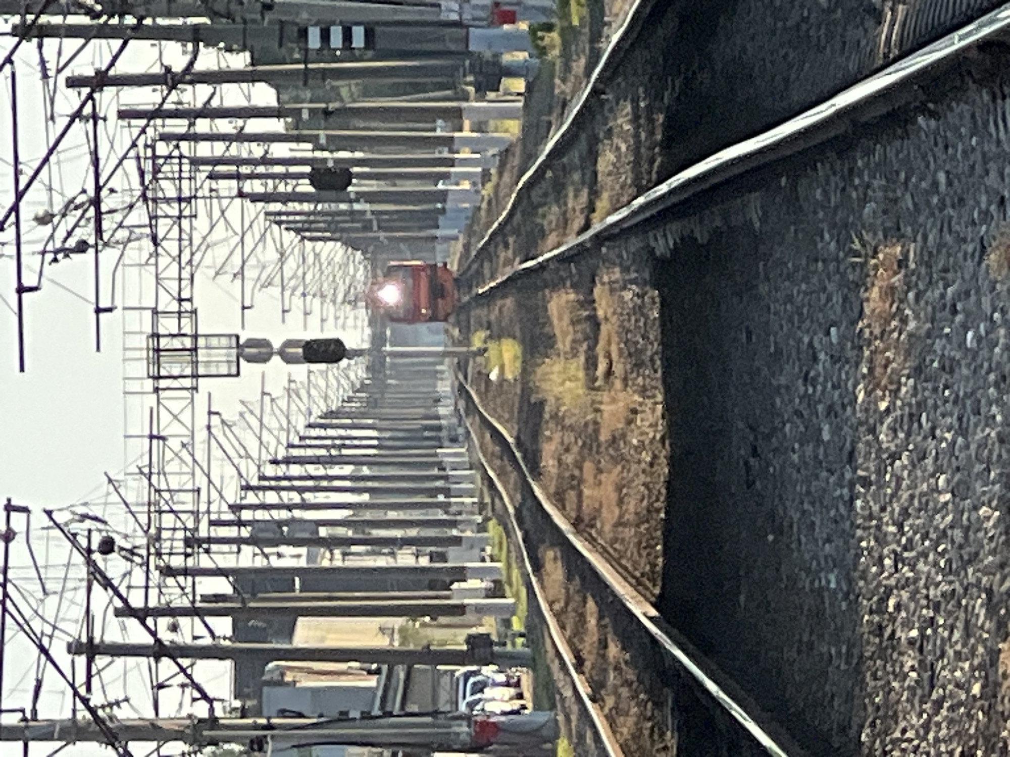 This image shows a train approaching on railway tracks in an outdoor environment. The railway tracks are in the foreground, with gravel ballast. Overhead power lines and supporting poles are visible along the tracks. In the distance, a red train with its headlight on is approaching. To the left of the tracks, a building with windows and cars parked nearby are visible. The scene appears to be in an industrial or transportation hub area. The lighting suggests it is daytime, possibly morning or late afternoon, given the long shadows and the quality of light. There are no people clearly visible in the image. The overall impression is one of an active railway line. The image does not contain any specific details that would allow for pinpointing the exact location within the city of Krasnodar, Russia, beyond it being a railway setting within that city.