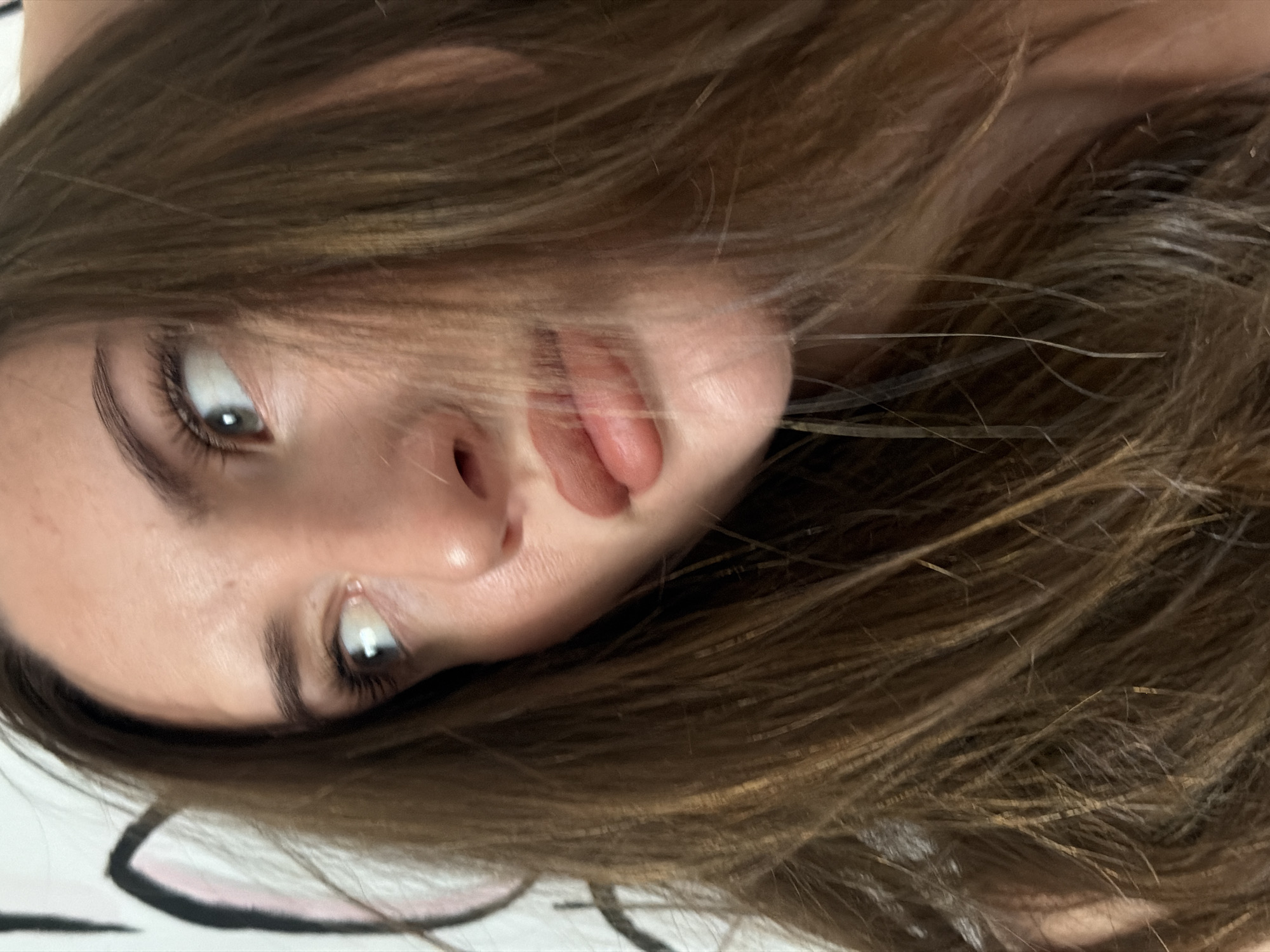 A close-up, low-angle, medium shot shows a young woman's face from the nose up, with her hair obscuring parts of her face. She has light brown hair, brown eyes, and is wearing light brown lipstick. She is looking to the right of the frame, with her mouth slightly open. The background is a white wall with a black and pink drawing of a cartoon character. The location of the image is not specified, and there are no details about the city of Sharm El Sheikh, Egypt.