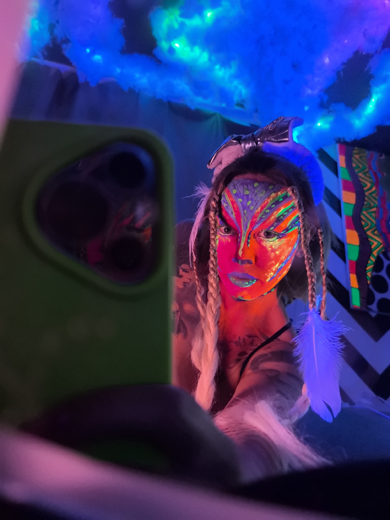 This vivid portrait captures a person with elaborate, glow-in-the-dark face paint under UV light. The individual's face is adorned with intricate neon patterns in vibrant shades of orange, pink, green, and blue, creating a distinctive, artistic look. Blonde hair is styled in braids, featuring a white feather accessory that glows blue. Tattoos are visible on their upper chest and shoulder. A dark, shiny headpiece or bow is worn on top of the head.
In the foreground, a bright green smartphone case, with its camera lenses reflecting the scene's colors, partially obstructs the view on the left, indicating the person is likely taking a selfie or engaging with the device.
The scene is an indoor environment, immersed in the dark, otherworldly ambiance of blacklight. Above, fluffy, cloud-like structures glow with intense blue and green light. On the right, a colorful tapestry with geometric patterns and tribal-like designs, including neon stripes and zigzags, adds to the vibrant setting. The strong UV lighting suggests a nighttime event, party, or artistic performance. The overall mood is energetic and artistic. No text is visible. The provided location context is Noviy, Russia.