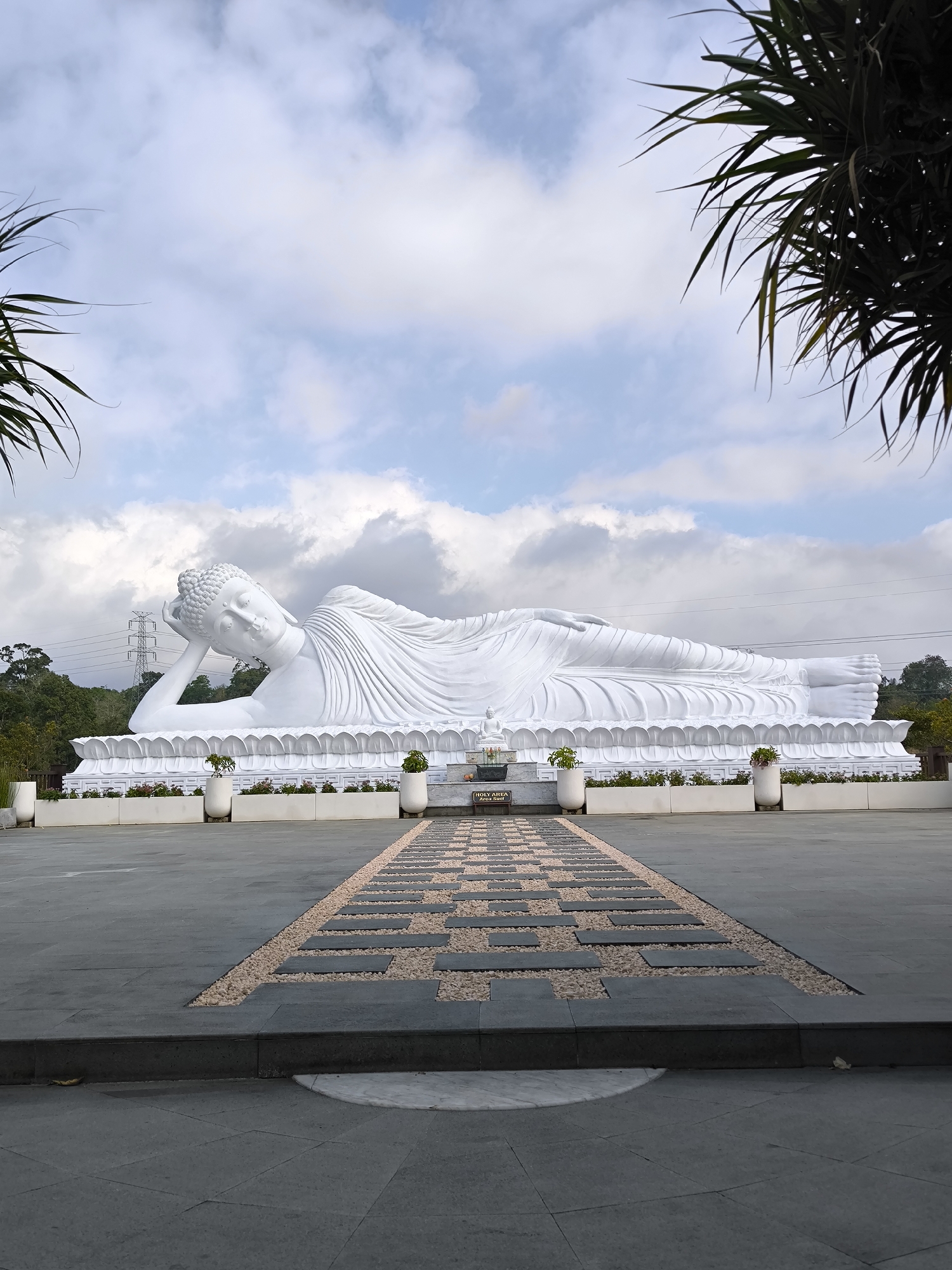 The image shows a large, white, reclining Buddha statue. The statue is positioned outdoors in what appears to be a temple or religious site. The statue is resting on a raised platform decorated with lotus petals. In front of the statue, there is a paved pathway with a decorative pattern of stepping stones and gravel. Two white planters with small green plants are placed on either side of the statue's base. In the background, there are trees and power lines. The sky is partly cloudy. The scene is located in Pupuan, Indonesia.