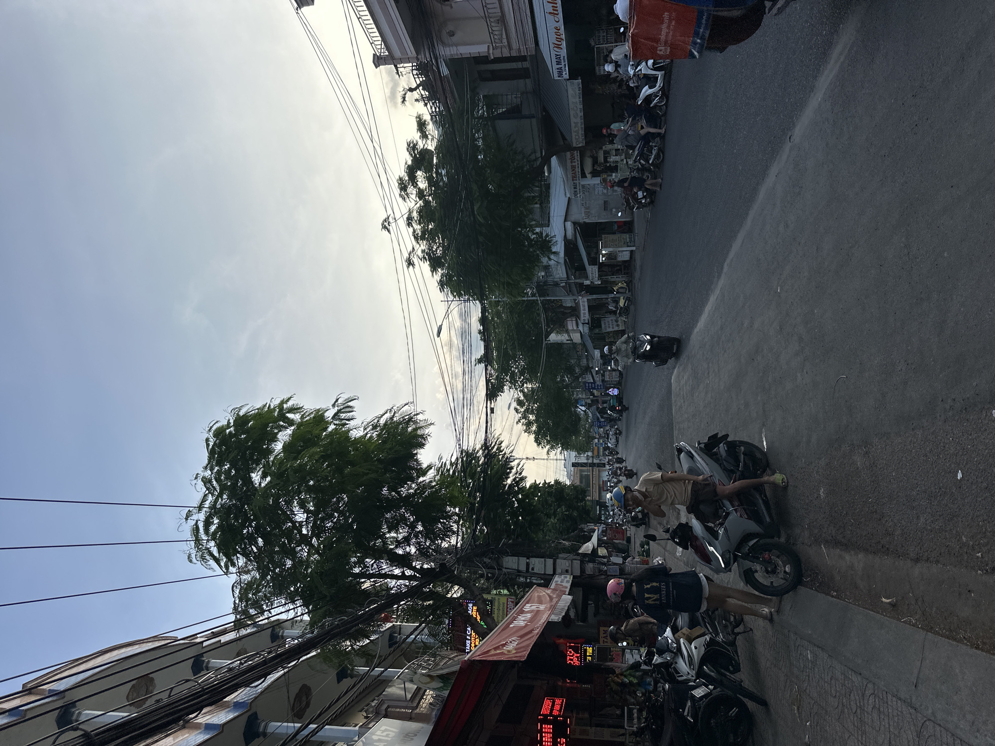 The image depicts a street scene in Bac Nha Trang Ward, Vietnam, during what appears to be late afternoon or early evening, given the lighting. The street is lined with buildings on the left and right sides, with a prominent presence of trees and a dense network of overhead electrical wires. A significant number of motorcycles are parked along the sidewalk on the left and are also in use on the street.
On the left side of the street, a commercial building with signs is visible, including one with red LED lights displaying text in what appears to be Vietnamese. Two individuals are standing on the sidewalk near a parked motorcycle. One, wearing a dark t-shirt with "NY YANKEES" printed on it and a pink helmet, is facing away from the camera. The other, wearing a light-colored short-sleeved shirt and dark shorts, with a blue helmet, is facing towards the camera and appears to be speaking.
The street itself is paved and appears to be a two-way road with traffic moving in both directions, primarily composed of motorcycles. On the right side of the street, more buildings are visible, some with awnings. A motorcycle with a large red box on the back, possibly for delivery, is parked on the right side of the road. The sky above is partly cloudy, with a gradient of blue and lighter tones, suggesting the sun is low on the horizon. The overall impression is a busy urban environment characterized by motorcycle-centric transportation and a dense overhead infrastructure of wires.