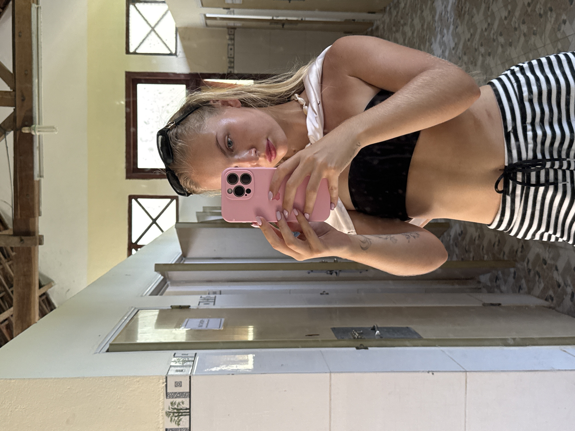 A young woman is taking a selfie in what appears to be a bathroom. She has blonde hair and is wearing sunglasses perched on her head, a black bikini top, and black and white striped shorts. She is holding a pink smartphone with a case in her right hand to take the photo. Her left arm is bent and resting on her hip.
The background shows a tiled wall and several doors, suggesting a communal area like a restroom. The lighting appears to be natural, possibly from windows visible in the reflection, indicating it is daytime. The overall mood seems casual and relaxed, likely a vacation setting. There is no visible text in the image.