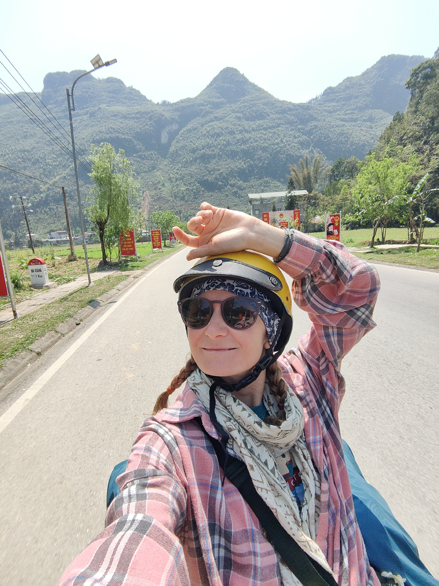 A woman wearing a yellow helmet and sunglasses is on a motorbike on a paved road. She is wearing a pink and grey plaid shirt and a patterned scarf. She has her hand on her helmet. The background shows mountains, trees, a power pole with a streetlight, signs, and other greenery. The road appears to be in good condition. A sign reads "Km 118, Ha Giang 113 Km". The photo was taken near Phố Bảng Commune, Vietnam.