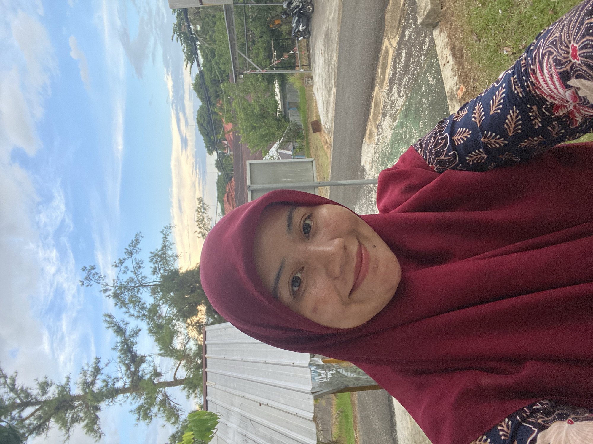 A mid-shot, eye-level, outdoor selfie captures a young woman wearing a maroon hijab and a patterned long-sleeved shirt. She is smiling with her head tilted slightly to her left. Her skin has some slight redness on her cheeks and forehead. In the background, a corrugated metal roof of a building is visible on the left, with trees and a cloudy blue sky overhead. To the right, there is a paved area with a few parked motorcycles and some green grass. The setting appears to be a residential or communal outdoor space within the city of Banjarbaru, Indonesia, possibly during the late afternoon or early evening given the soft lighting.
