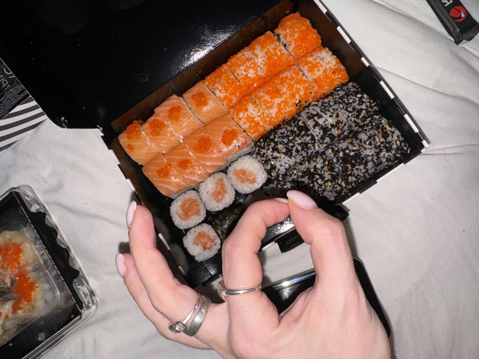 The image presents an overhead view of two sushi containers resting on a light-colored, textured fabric surface, likely inside a residence in Moscow, Russia. A fair-skinned human left hand with manicured light pink fingernails is positioned on the edge of the primary, open black sushi box. The hand wears two silver-toned bands on the index finger and a single silver-toned band on the ring finger.
The main black sushi box contains four distinct types of sushi rolls. From left to right: four maki rolls with visible salmon filling, followed by six rolls topped with slices of salmon and orange fish roe (tobiko). To the right of these are eight rolls coated in a mixture of black and white sesame seeds, and furthest right, eight rolls fully covered in orange tobiko. A second, partially visible clear plastic container in the upper left also holds sushi rolls, identifiable by orange tobiko. Additional elements in the periphery include a piece of black and white striped packaging with indistinct Russian text and a small black packet with a red logo, positioned in the lower right corner. The lighting suggests an indoor setting, possibly evening.