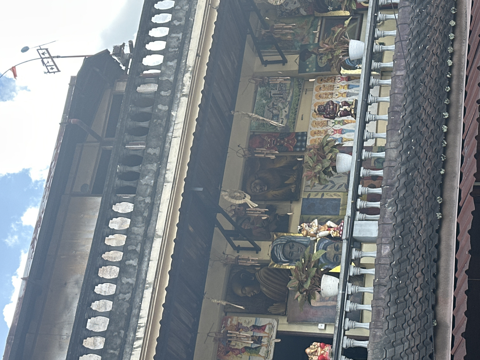 The image shows the exterior of a building in Ubud District, Indonesia, likely a shop or gallery displaying various artworks and religious figurines. The building has multiple levels, with balconies adorned with ornate railings and decorative pillars. Several paintings depicting religious figures, animals, and landscapes are hung on the walls. Various statues, including what appears to be a depiction of Ganesha, and numerous wind chimes are also visible. Potted plants are strategically placed on the balconies. The roof is covered with traditional dark tiles, and in the background, the sky with clouds and a weather vane can be seen. The overall impression is a vibrant and culturally rich display of local artistry.