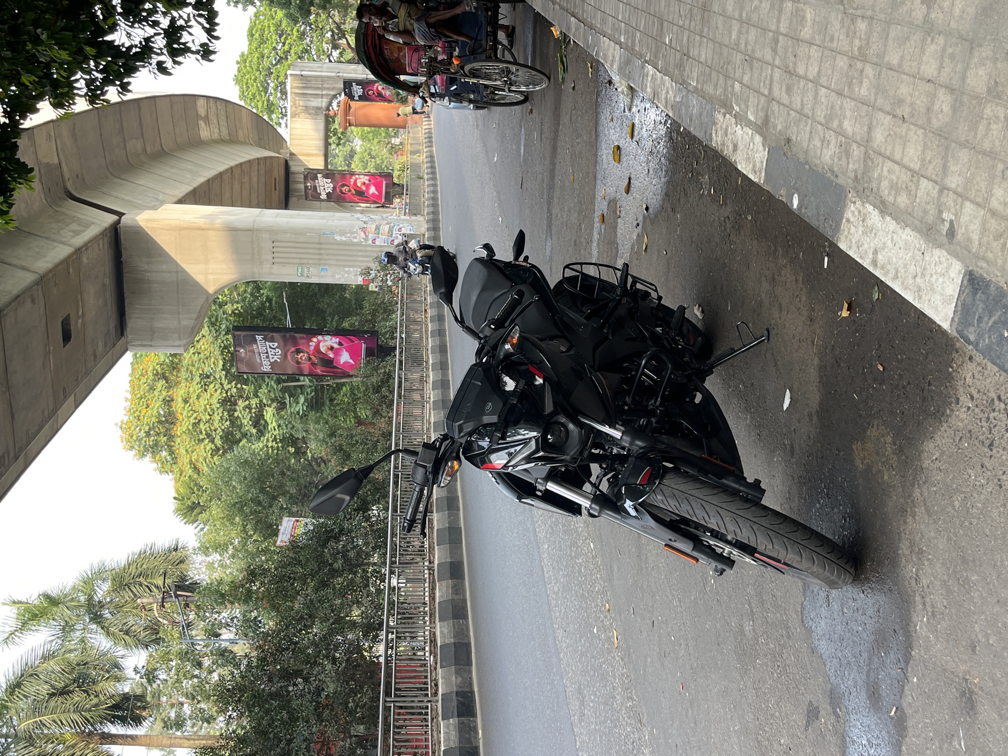 This outdoor street scene in Dhaka, Bangladesh, captures a black motorcycle parked on the side of an asphalt road. To the right of the road, a tiled sidewalk runs along. The ground has scattered leaves and wet patches, suggesting recent moisture or spilled liquid.
On the left side of the road, lush green trees, including palm trees, line the area. Above the foliage and road, a massive, curving concrete elevated structure, likely a flyover or metro rail viaduct, dominates the background. Sunlight casts clear shadows on its underside, indicating a bright daytime.
Further down the road, a traditional rickshaw with a person inside is visible, along with another motorcycle. The pillars supporting the elevated structure display several advertisements. These vibrant posters, predominantly pink and red, feature images of people and Bengali text. "বিকাশ" (bKash), a mobile financial service, is clearly visible on these ads, along with phrases such as "বিকাশ অ্যাপে" (on bKash App). One concrete pillar also bears the numerical marking "P50-80."
The scene depicts a typical busy urban setting in daylight, characterized by local transportation, infrastructure, and commercial advertising.