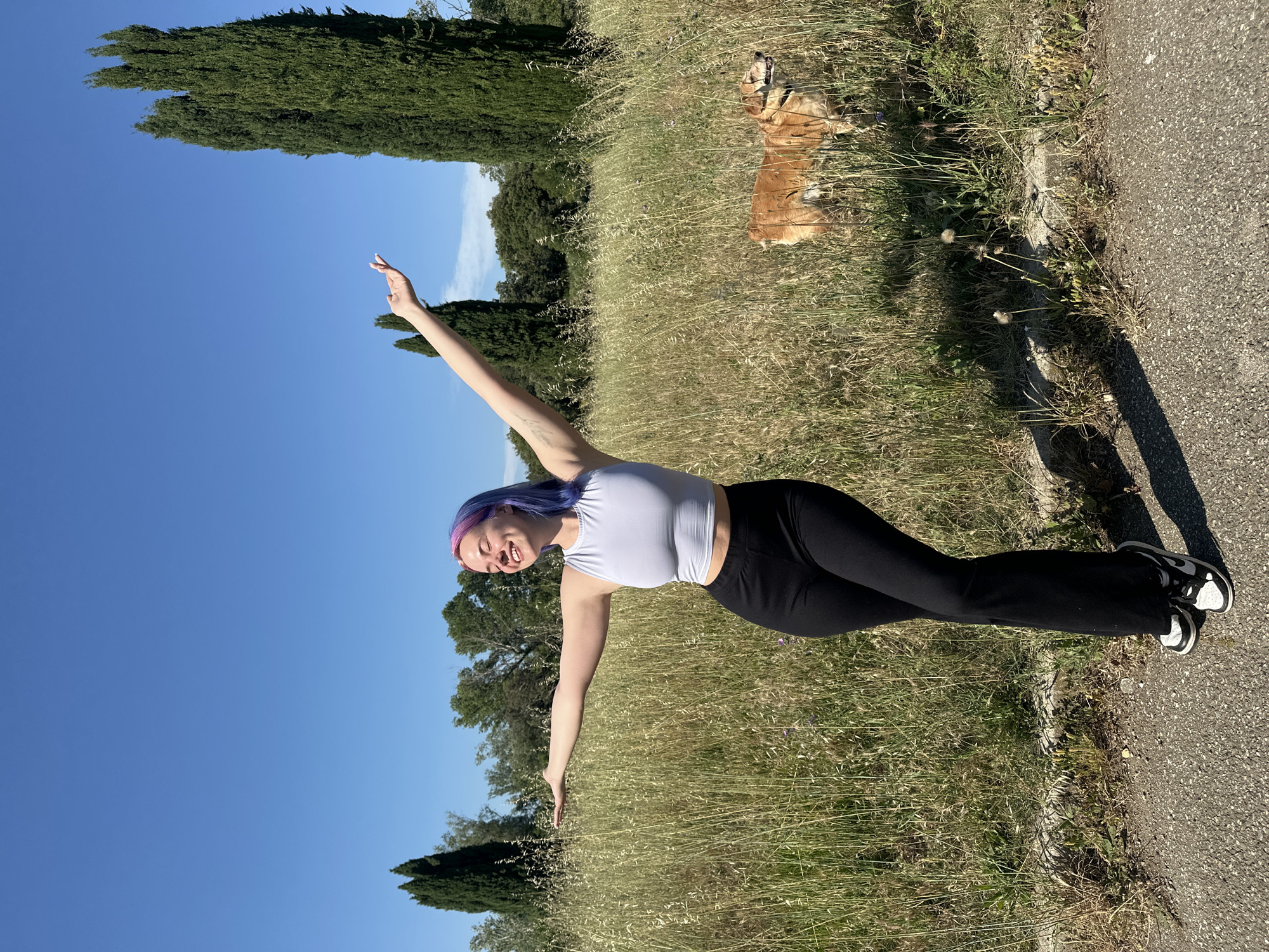 A young woman stands in a field of tall, dry grass in Caissargues, France, under a clear blue sky, with trees lining the horizon. She wears a white tank top and black pants, and her hair is dyed purple. She stands with her arms outstretched, smiling. A dog rests in the grass behind her. A paved pathway runs along the edge of the field, casting a shadow of the woman.