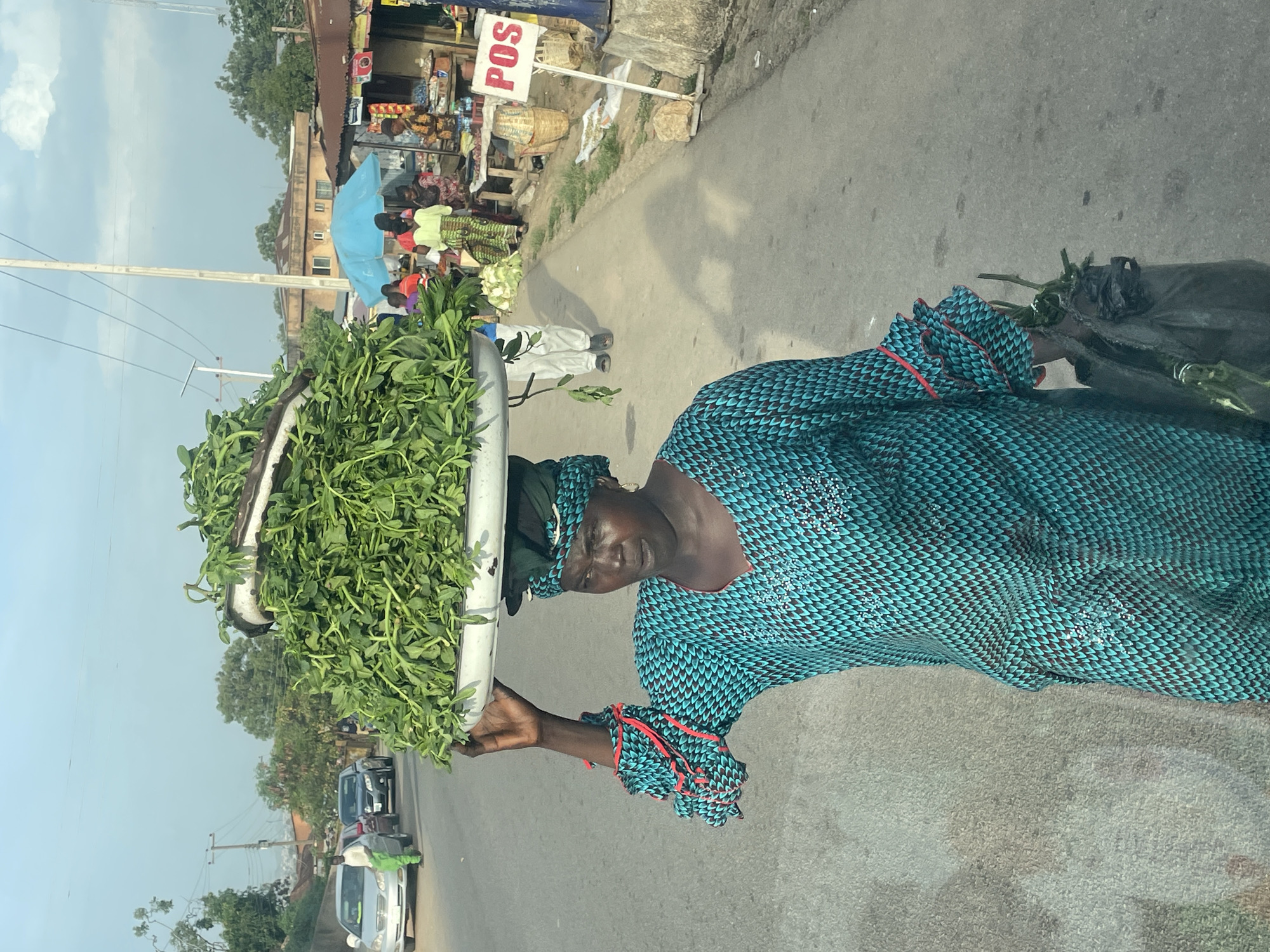 An African woman walks along a road in what appears to be the city of Jos, Nigeria. She carries a large, round, silver-colored tray on her head filled with leafy green vegetables. The woman wears a patterned blue and black dress with red and blue trim on the sleeves. She is walking away from a market area with shops and stalls, some shaded by a blue umbrella. A "POS" sign is visible near the market area. Cars are parked on the opposite side of the road. The sky is partly cloudy.