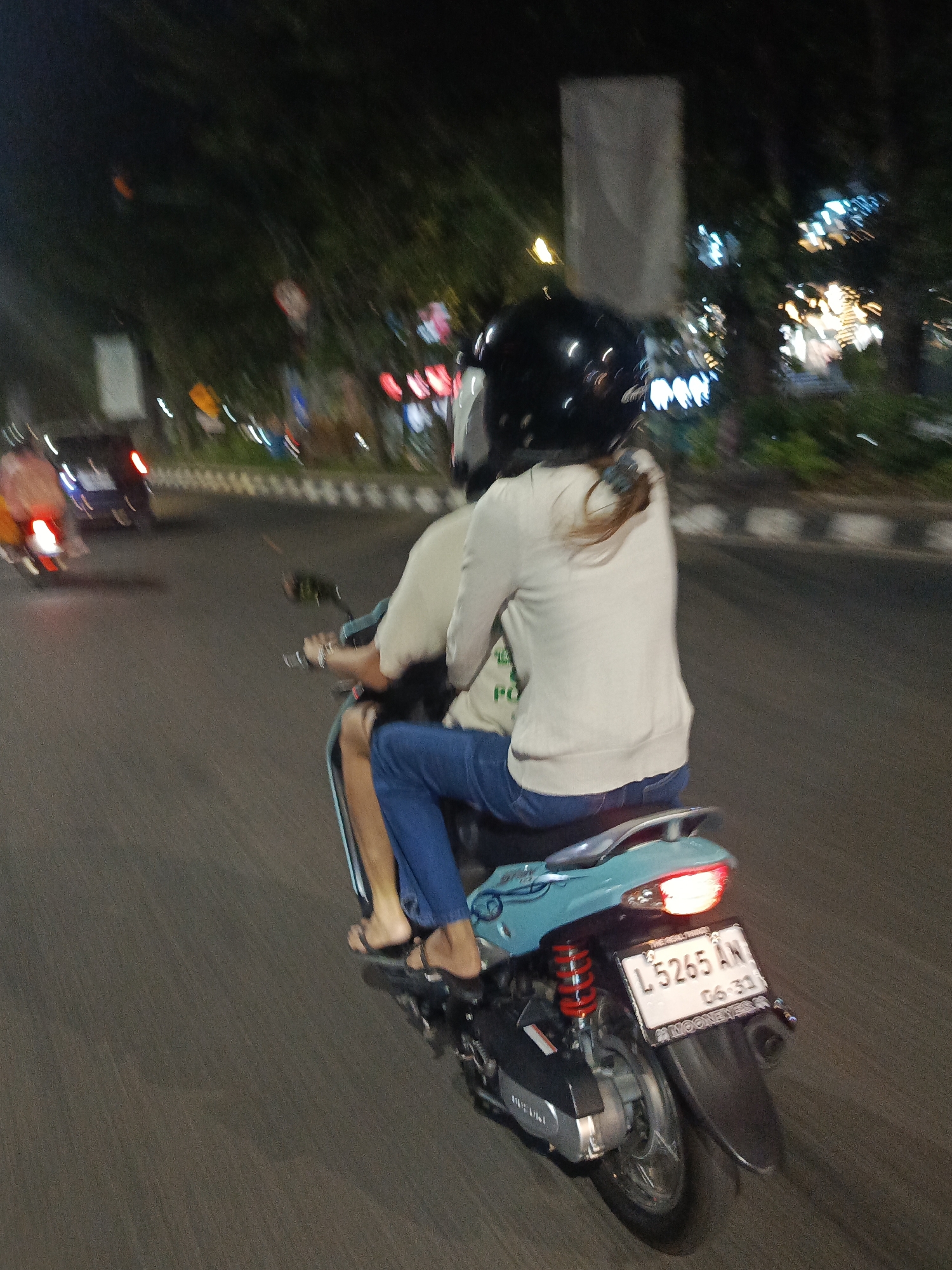 A rear view, night-time, motion-blurred photograph captures two individuals riding on a teal-colored scooter on an asphalt road in Surabaya, Indonesia. The front rider, wearing a black helmet, faces away from the camera. The back rider, also facing away, is wearing a light-colored long-sleeved shirt and blue jeans. Their feet are bare, wearing dark flip-flops. The scooter has a red taillight illuminated and a white license plate that reads "L 5265 AN" and "06 33". To the left of the scooter, a dark-colored car with illuminated taillights is in motion. The background is a blur of dark trees and streetlights. The scene appears to be on a city street with a pedestrian sidewalk visible in the middle ground.
