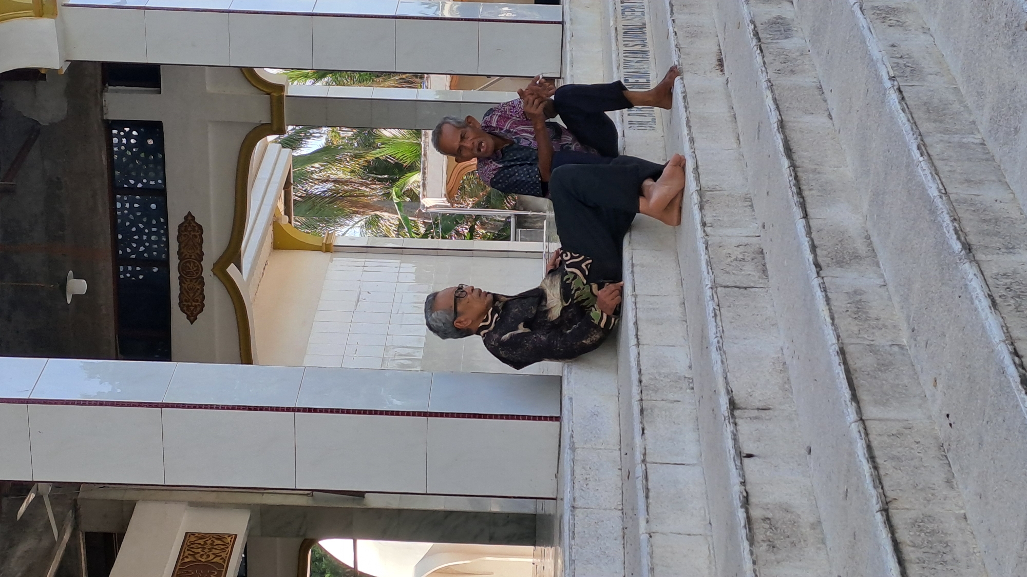 Two elderly men are sitting on the steps of a building, possibly a mosque given the tiled walls and decorative Islamic calligraphy visible on the building's exterior. The man in the foreground, wearing glasses and a dark patterned shirt, is facing to the right, looking down with a thoughtful expression. The man behind him, with grey hair and wearing a reddish-purple patterned shirt, is looking to his left and appears to be speaking or gesturing. Both men are barefoot and dressed in traditional Indonesian attire. The steps are made of rough-textured stone. Palm trees and other greenery are visible in the background, suggesting a tropical climate. The lighting indicates it is daytime, likely late morning or early afternoon, with bright sunlight casting shadows. An inscription is visible on the side of the steps, though it is difficult to read clearly.