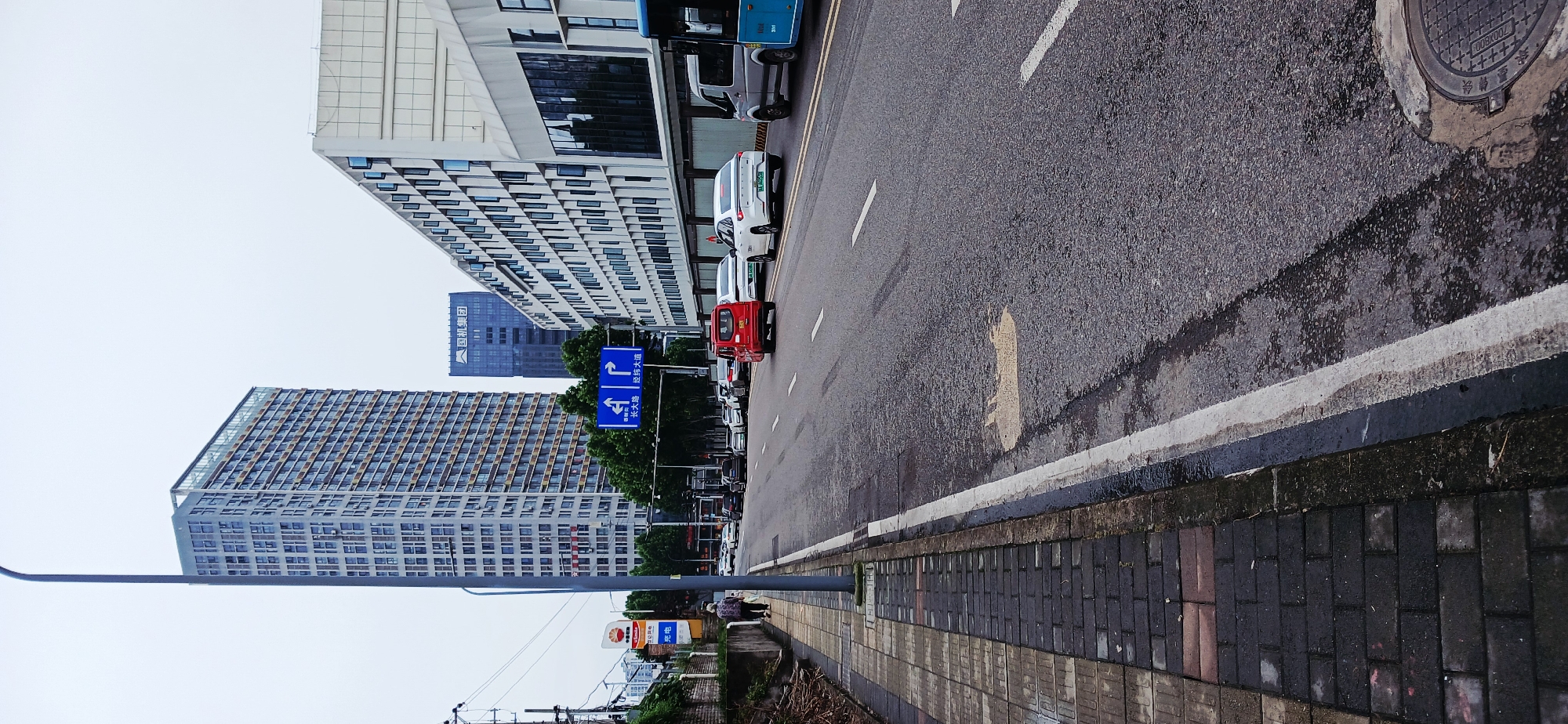 The photograph depicts a street scene in Jiulongpo District, Chongqing, China. A multi-lane road stretches into the distance, lined with parked vehicles. A tall, modern building with a grid-like facade dominates the left side of the frame. Another high-rise building is visible behind it. Street signs are posted along the road. A sidewalk runs along the right side of the road, paved with dark bricks. A street lamp extends over the sidewalk. The sky is overcast. Two people walk on the sidewalk in the middle distance. A manhole cover is in the upper right corner of the image.
