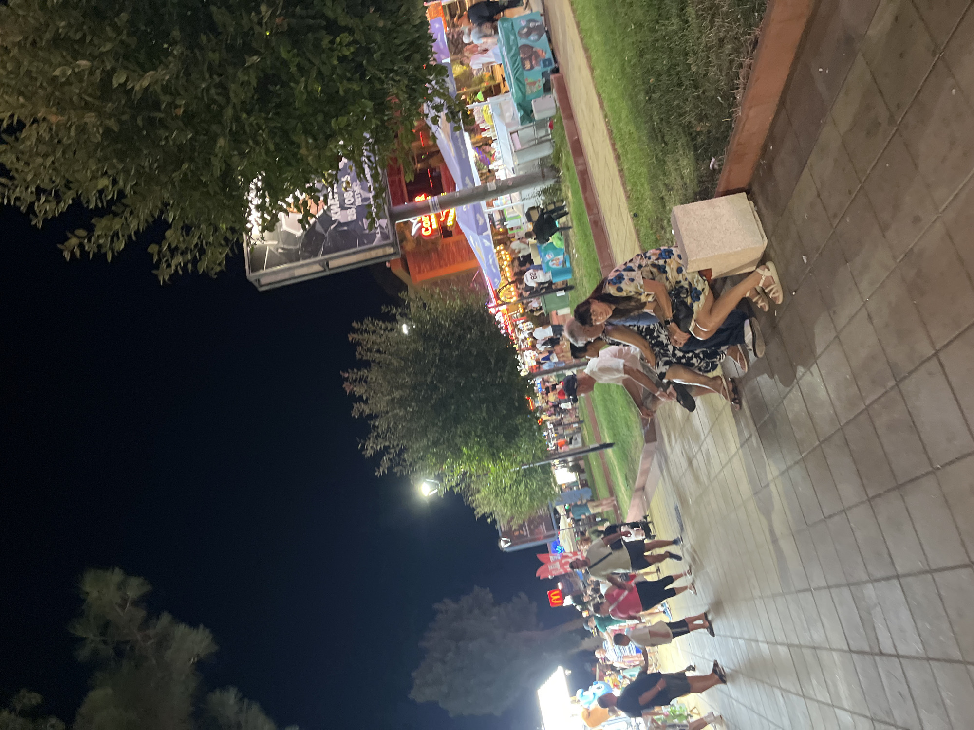 In the city of Nesebar, Bulgaria, a nighttime scene unfolds along a bustling promenade. The image captures a segment of a street lined with various establishments, indicated by illuminated signs and awnings. People are seen strolling along the paved walkway and sitting on a low concrete bench in the foreground. A group of individuals, including men, women, and children, are dispersed throughout the scene, some walking in pairs or small groups, others seated. The environment is characterized by trees, streetlights, and the vibrant glow of commercial activity. The overall atmosphere suggests a lively evening, typical of a popular tourist destination.