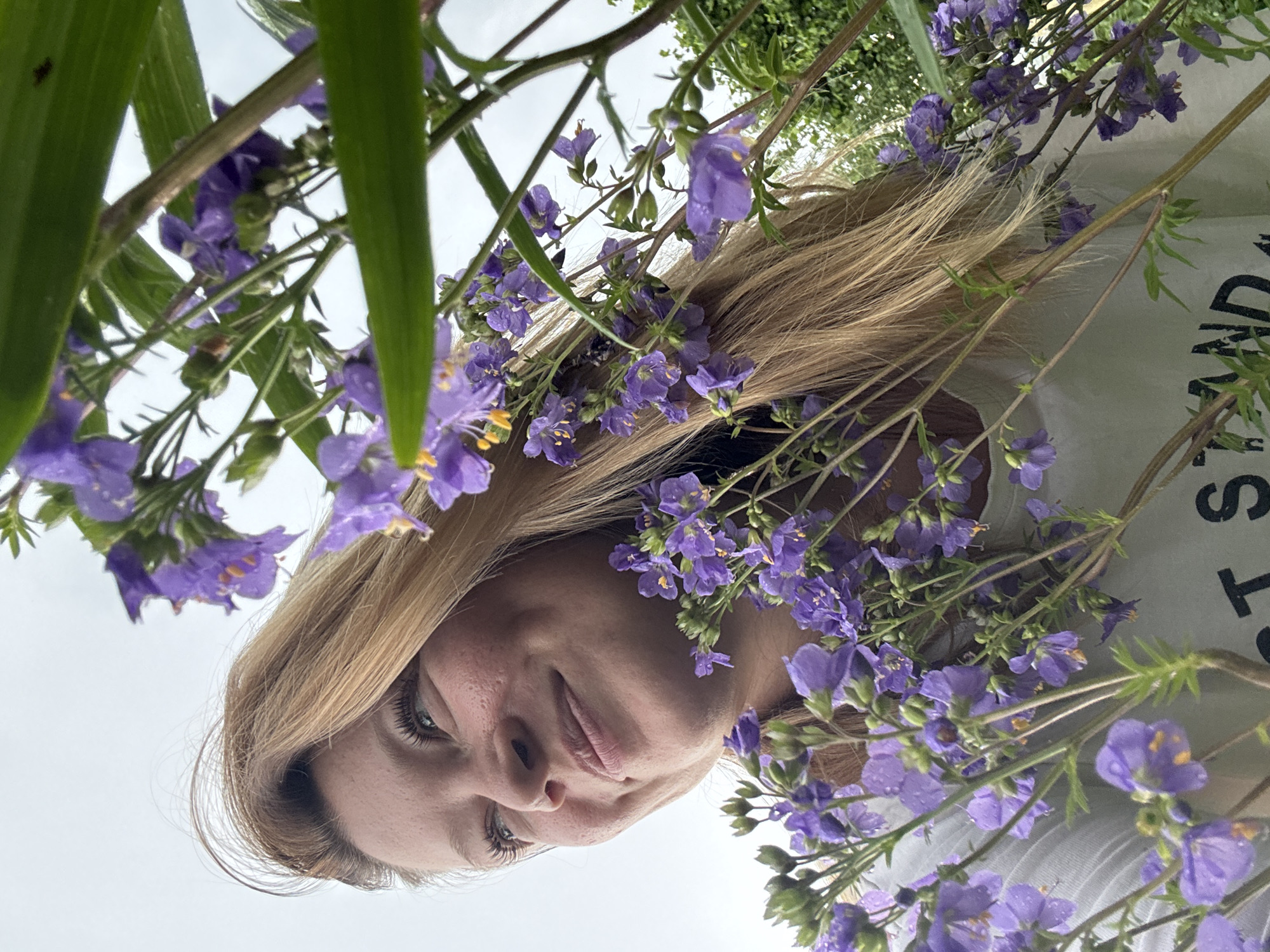 A young Caucasian woman with blonde hair and fair skin is pictured outdoors, with a shallow depth of field, obscuring the background. She is positioned in the lower left quadrant of the image, looking to her left with a slight smile. Her hair is long and blonde, falling around her face and shoulders. She is wearing a white t-shirt with black text visible on the front, part of which reads "ST STAND". The woman is surrounded by a profusion of small, purple flowers with yellow centers, which are in focus and occupy the majority of the frame. Some of the flowers and leaves have water droplets on them, suggesting recent rain or dew. The overall lighting is soft and diffused, indicating an overcast sky. There is no specific indication of the city of Vassyata, Russia in the image.